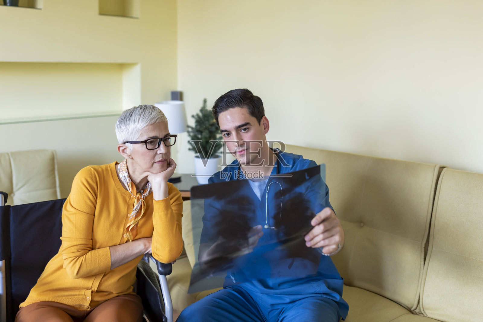 身穿蓝色制服的年轻男医生正在拜访一位坐在轮椅上的年长妇女，并在x光图像上诊断。照片摄影图片