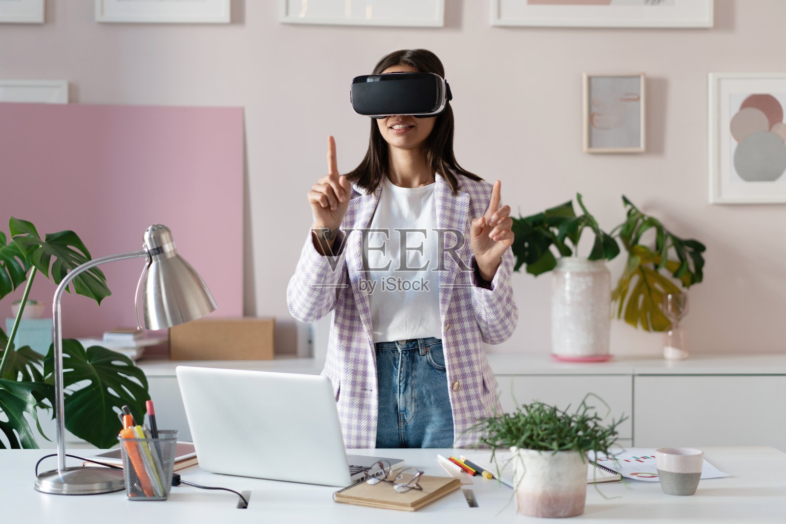 自信的年轻女子戴着虚拟现实头盔，站在办公室的工作地点，手指着空中照片摄影图片