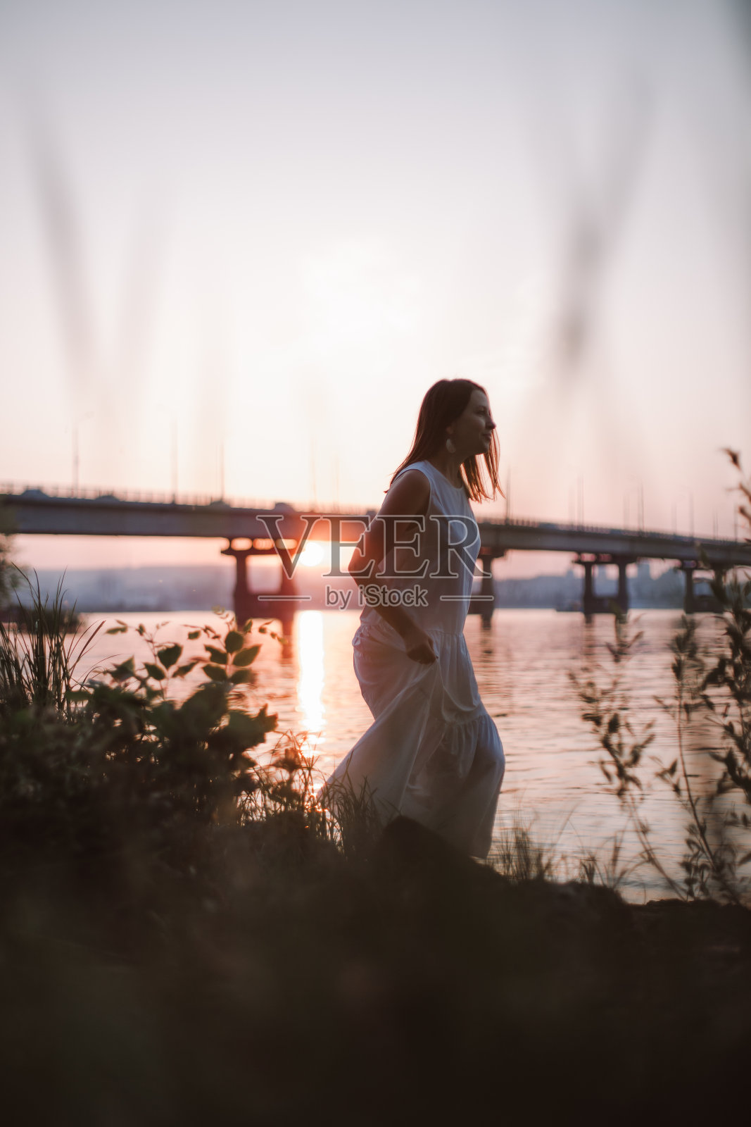 日落时分，妇女们沿着河岸散步。日落时分，一个身穿白色夏装的黑发女子沿着长满青草和树木的河岸散步照片摄影图片