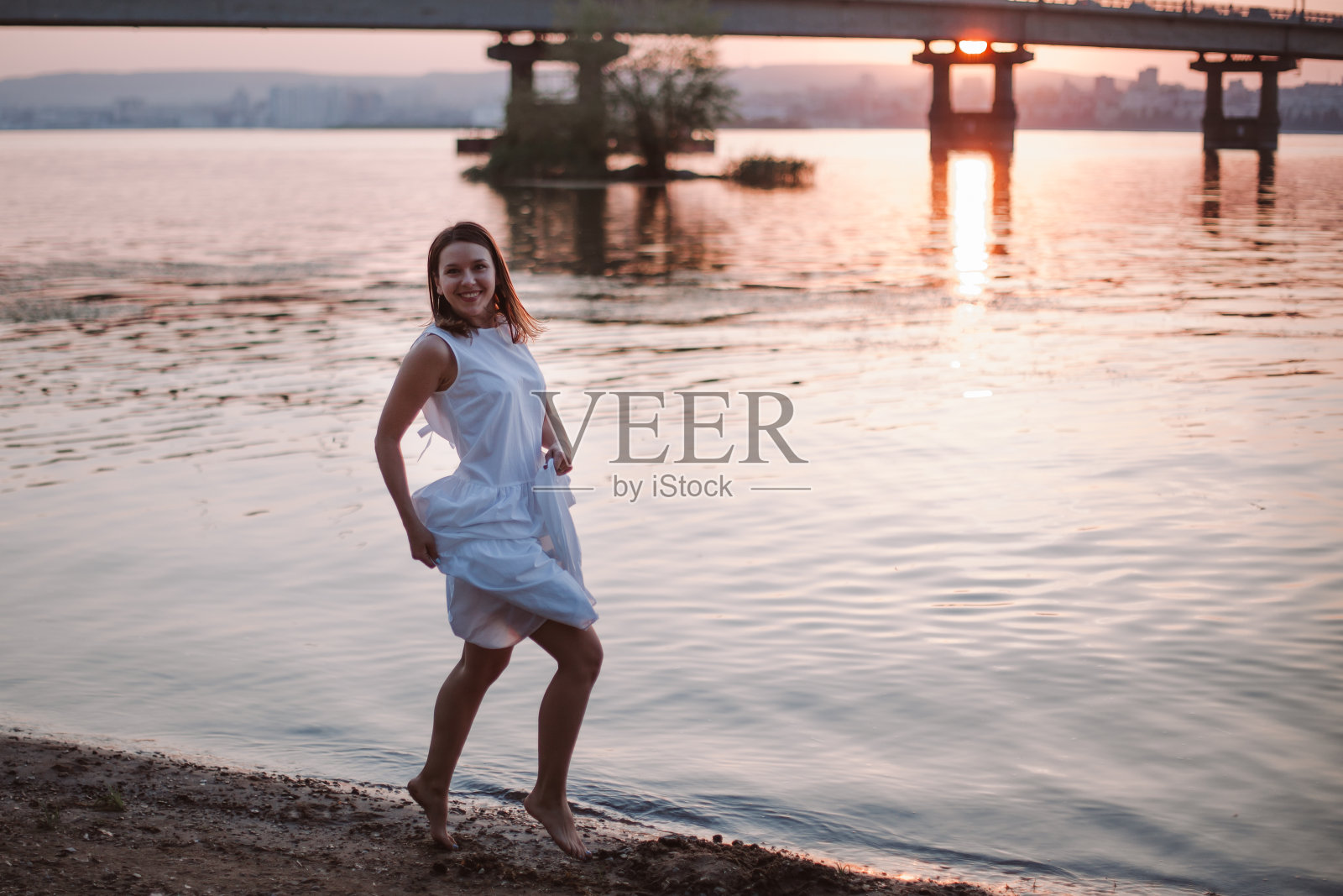 女人在日落时跳跃。一个笑着，快乐的女人穿着白色的夏装正在一个夏天的傍晚在河边的海滩上玩得很开心照片摄影图片