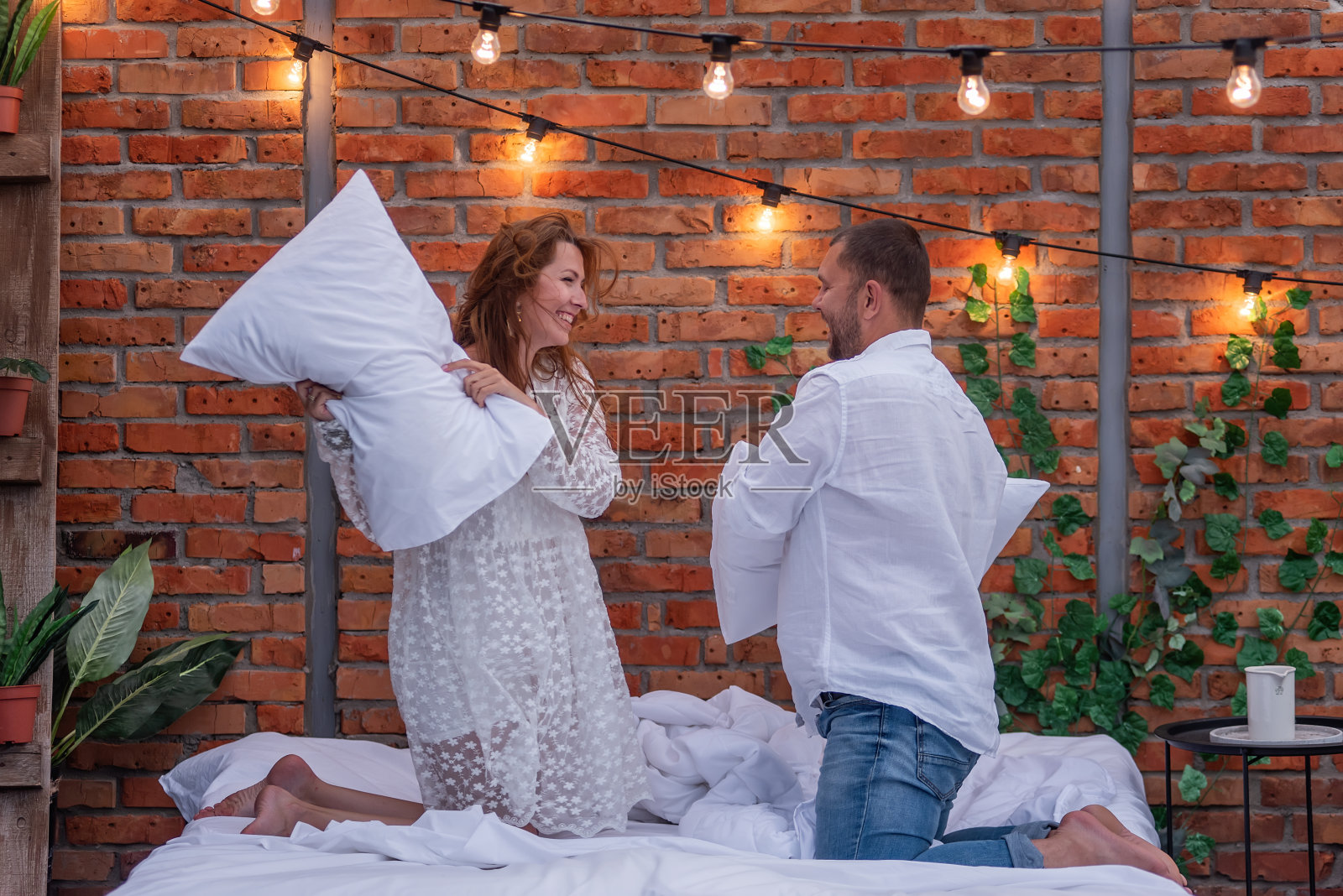 相爱的情侣在床上嬉戏，打枕头笑。美丽的女孩穿着白色的连衣裙，年轻人穿着牛仔裤，衬衫。以砖墙为背景，屋顶上有灯泡的花环照片摄影图片