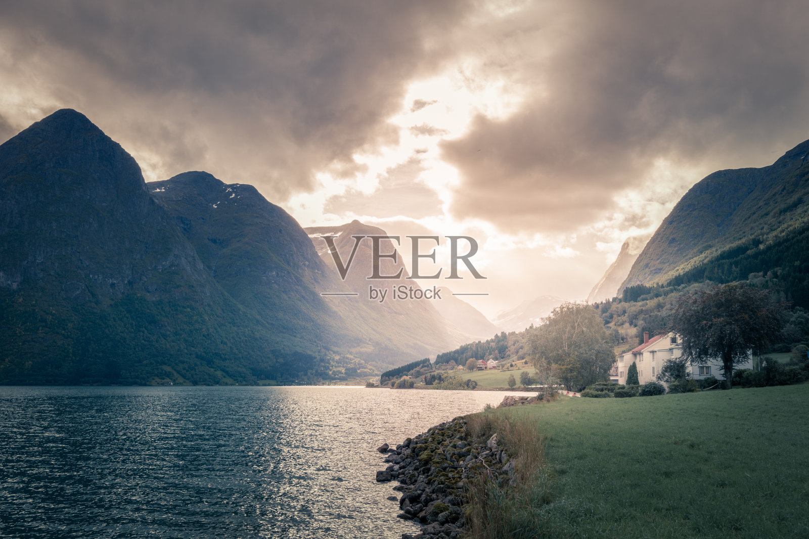 在挪威峡湾壮观的山脉之上，乌云和阳光穿过乌云。从海边看，附近有白色的斯堪的那维亚房子照片摄影图片