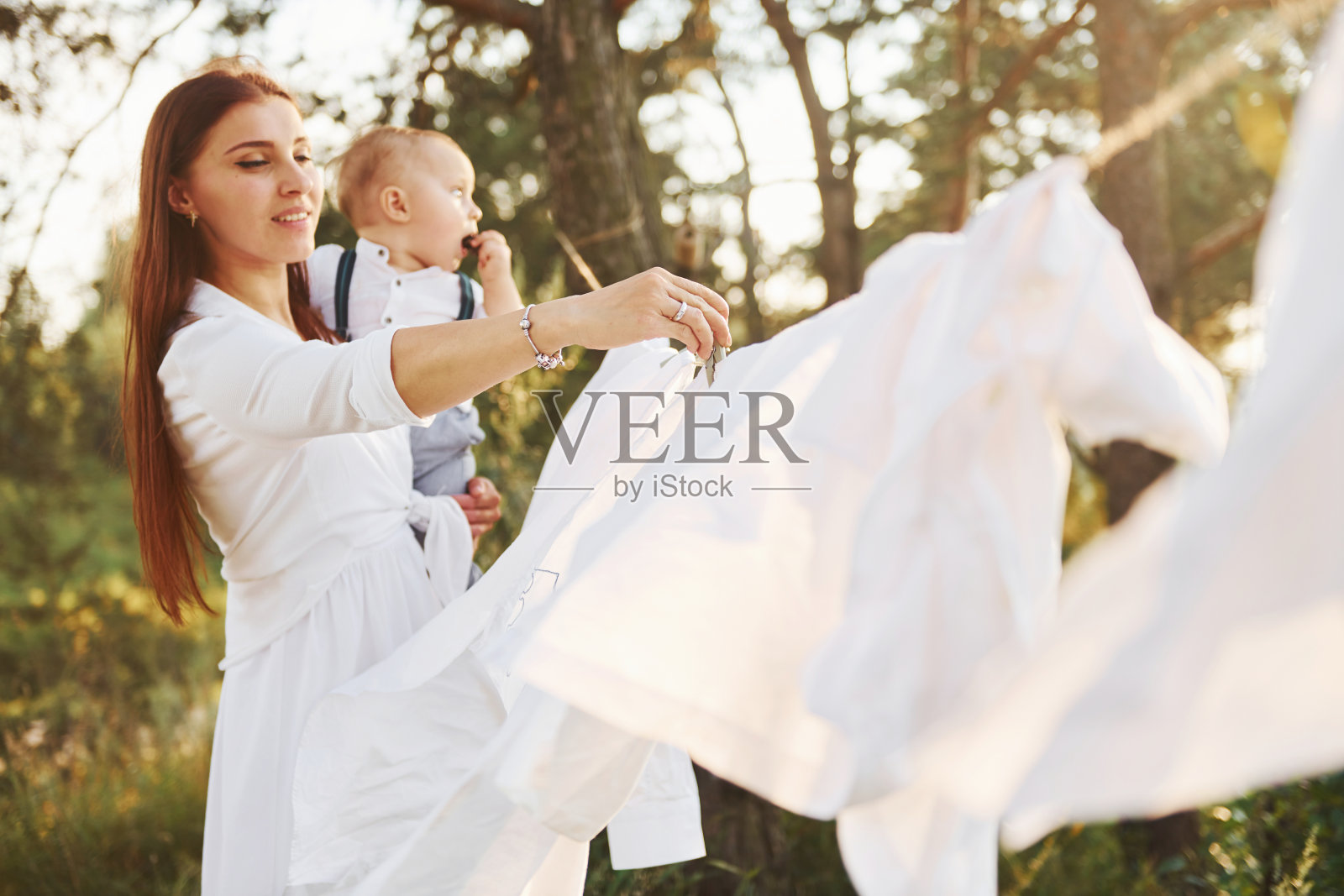 积极主妇用挂白衣服晾干。一位年轻的母亲带着她的小儿子在森林的外面。美丽的阳光照片摄影图片