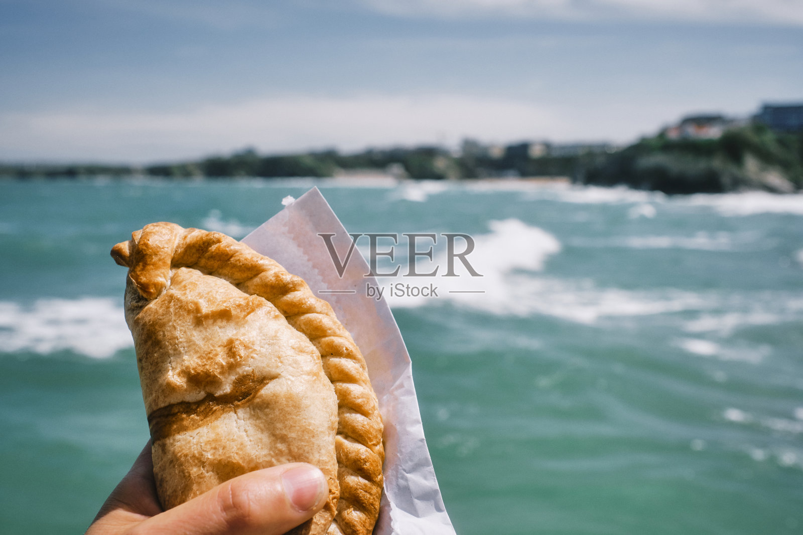 六月阳光明媚的纽基，一只手举着康沃尔馅饼在蓝色的海面上。照片摄影图片