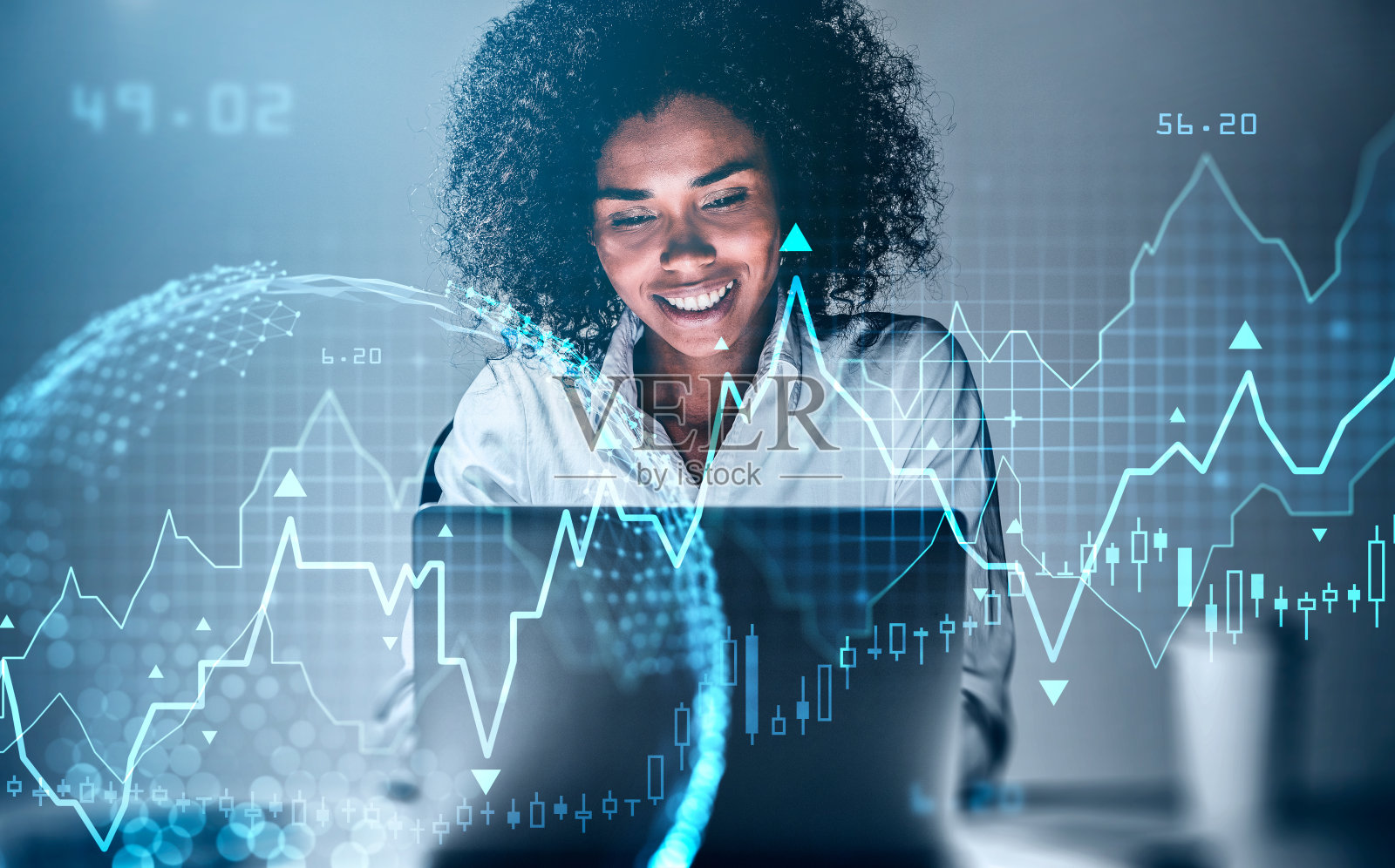 微笑的非裔美国女商人用笔记本电脑研究金融新闻。股票市场变化烛台，数据信息图标。地球投影。金融和技术的概念照片摄影图片