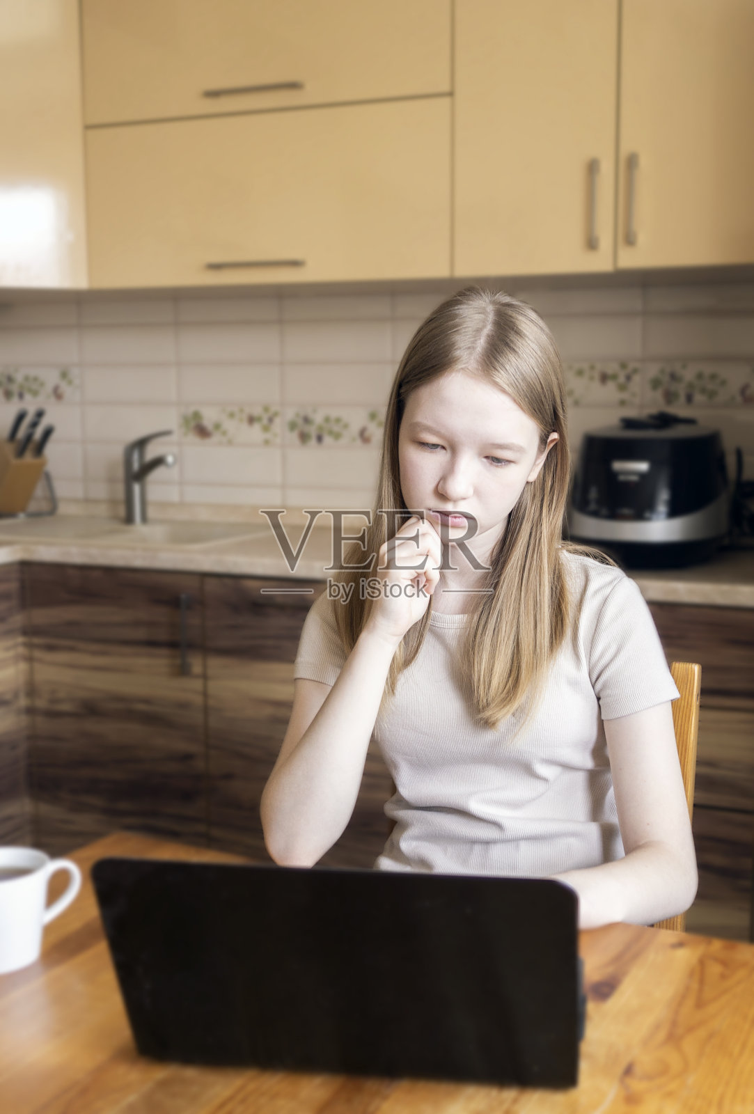 女孩看着笔记本电脑的屏幕。在家学习，看视频和网上聊天。一个十几岁的孩子在厨房。照片摄影图片