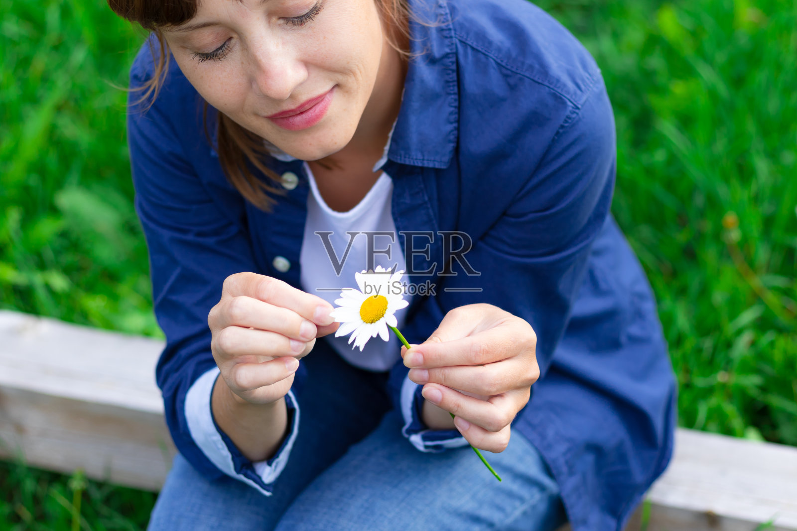 一个穿着蓝色衬衫的漂亮年轻女子正在草地上猜一朵甘菊。有选择性的重点照片摄影图片