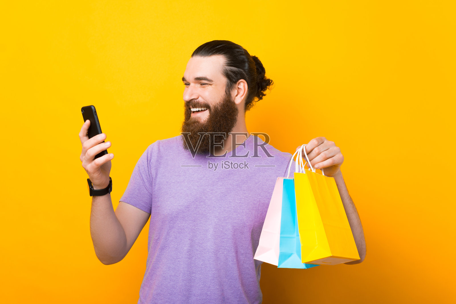 英俊的大胡子男人，拿着购物袋，使用手机，在黄色背景下微笑照片摄影图片