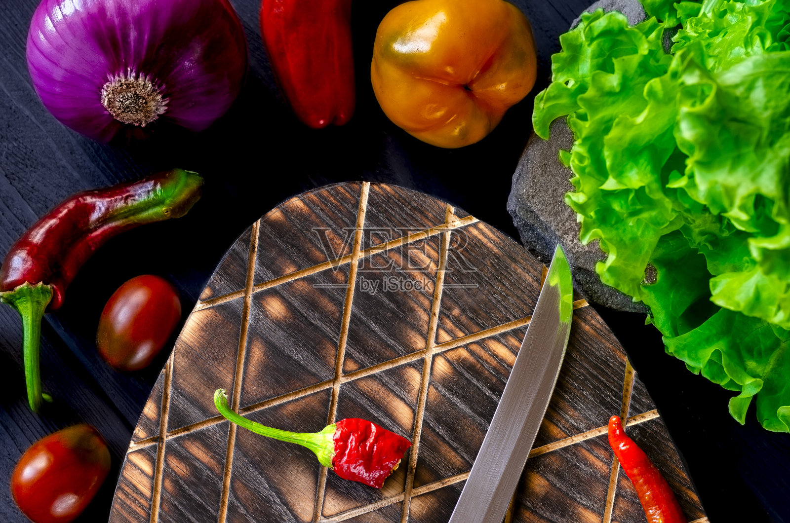 红色、黄色、绿色的辣椒、辣椒、蓝色的洋葱和绿色的莴苣放在一张黑色的木桌上，旁边的切菜板上放着青椒和一把刀照片摄影图片