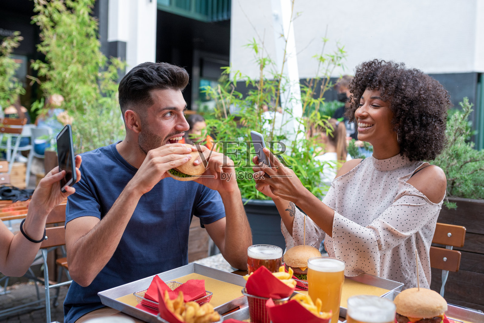 一群多民族的朋友在餐厅里玩得很开心，年轻的男人吃着汉堡包，非洲女人的朋友用她的手机给他拍照照片摄影图片