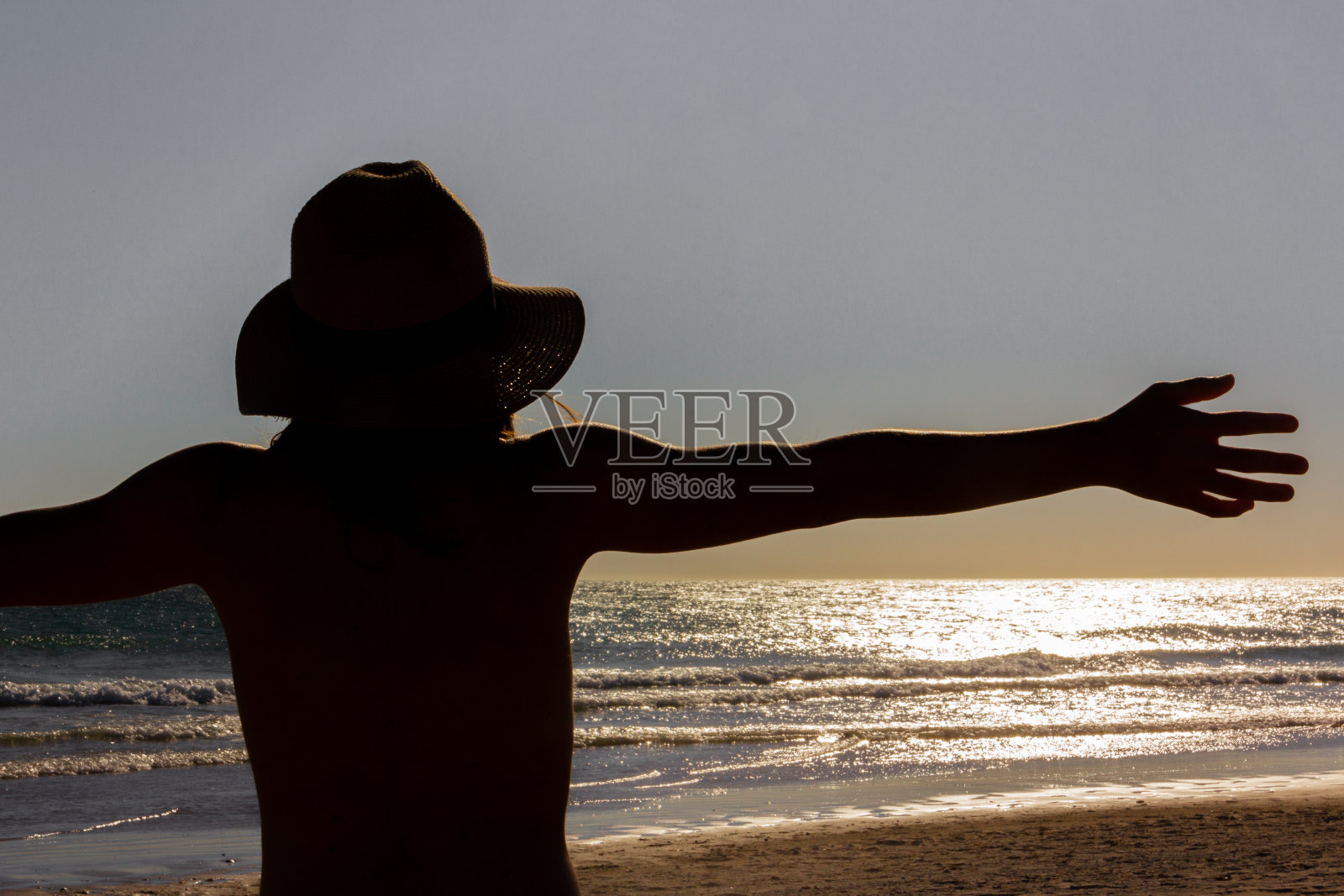 年轻的女孩戴着帽子，伸开双臂看着日落的大海。选择性focus.Backlighting。照片摄影图片