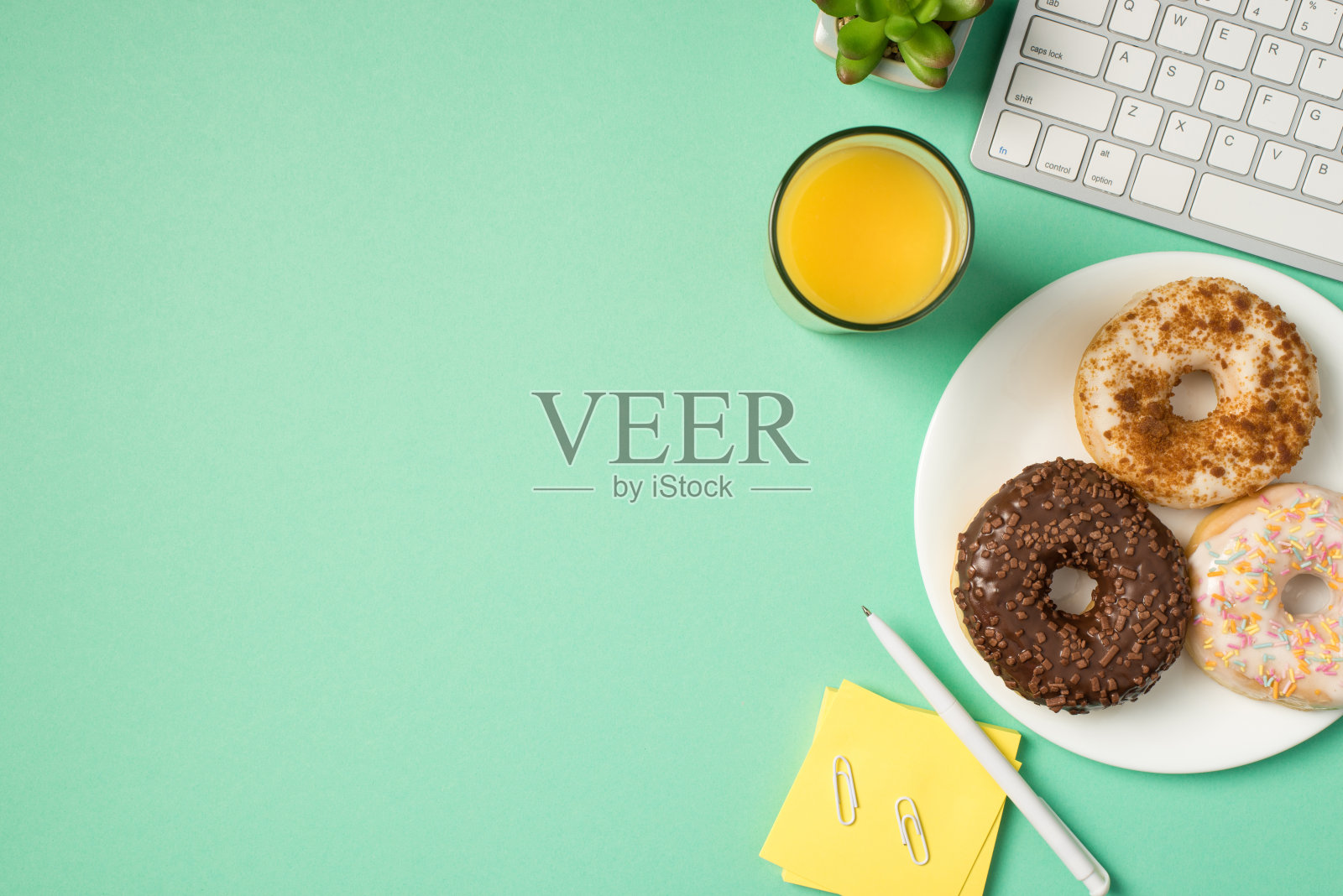 顶视图照片的工作场所键盘，笔夹，便签纸植物，果汁和三个甜甜圈在盘子上孤立的蓝绿色背景与空白的空间照片摄影图片