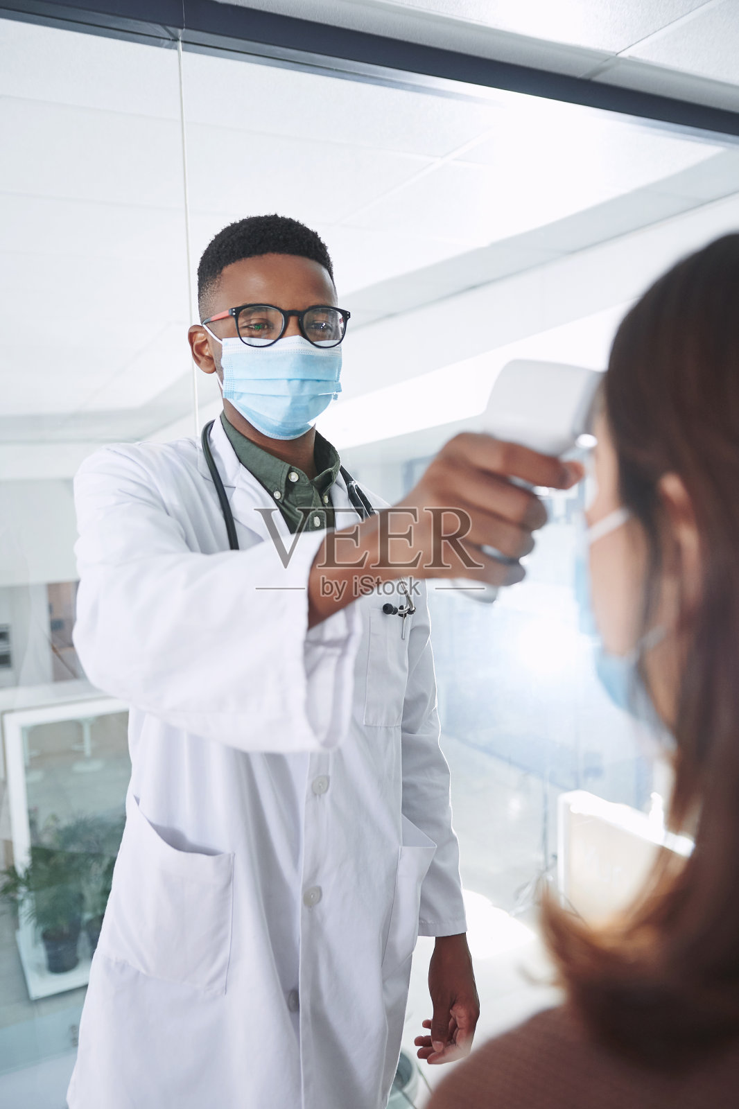 图为一名戴着口罩的年轻医生和病人一起站在诊所里给病人量体温照片摄影图片