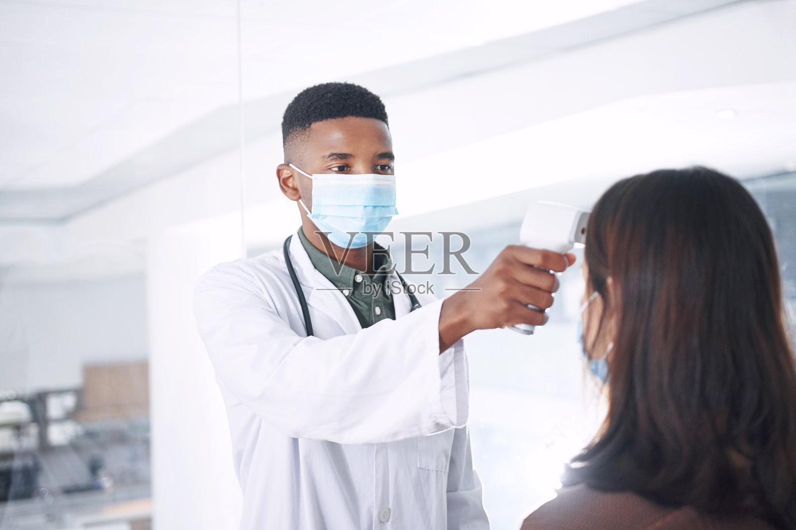 图为一名戴着口罩的年轻医生和病人一起站在诊所里给病人量体温照片摄影图片