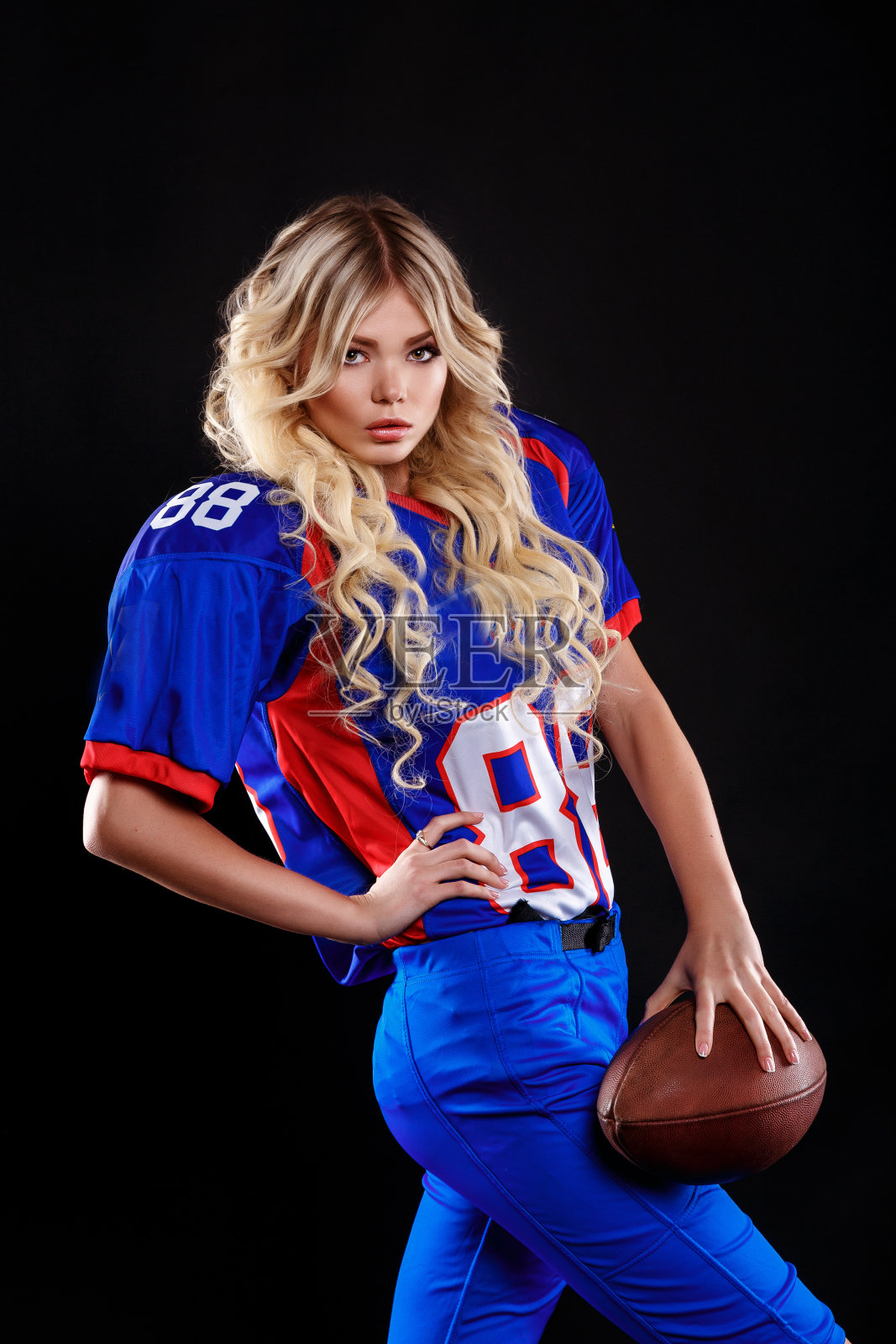 金发碧眼的运动员在黑色背景下扮演美式橄榄球女孩。漂亮的年轻女子穿着美式橄榄球帽，手里拿着球。美国女足球运动员的摄影照片摄影图片