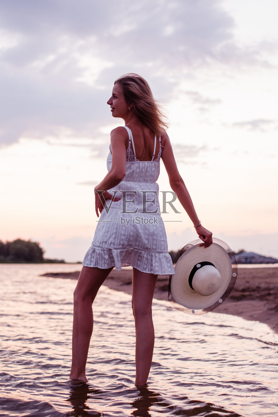 一个女人手里拿着一顶草帽。一个美丽快乐的金发女郎，穿着白色的夏装，挽着裙子的下摆，站在河边，直到日落照片摄影图片