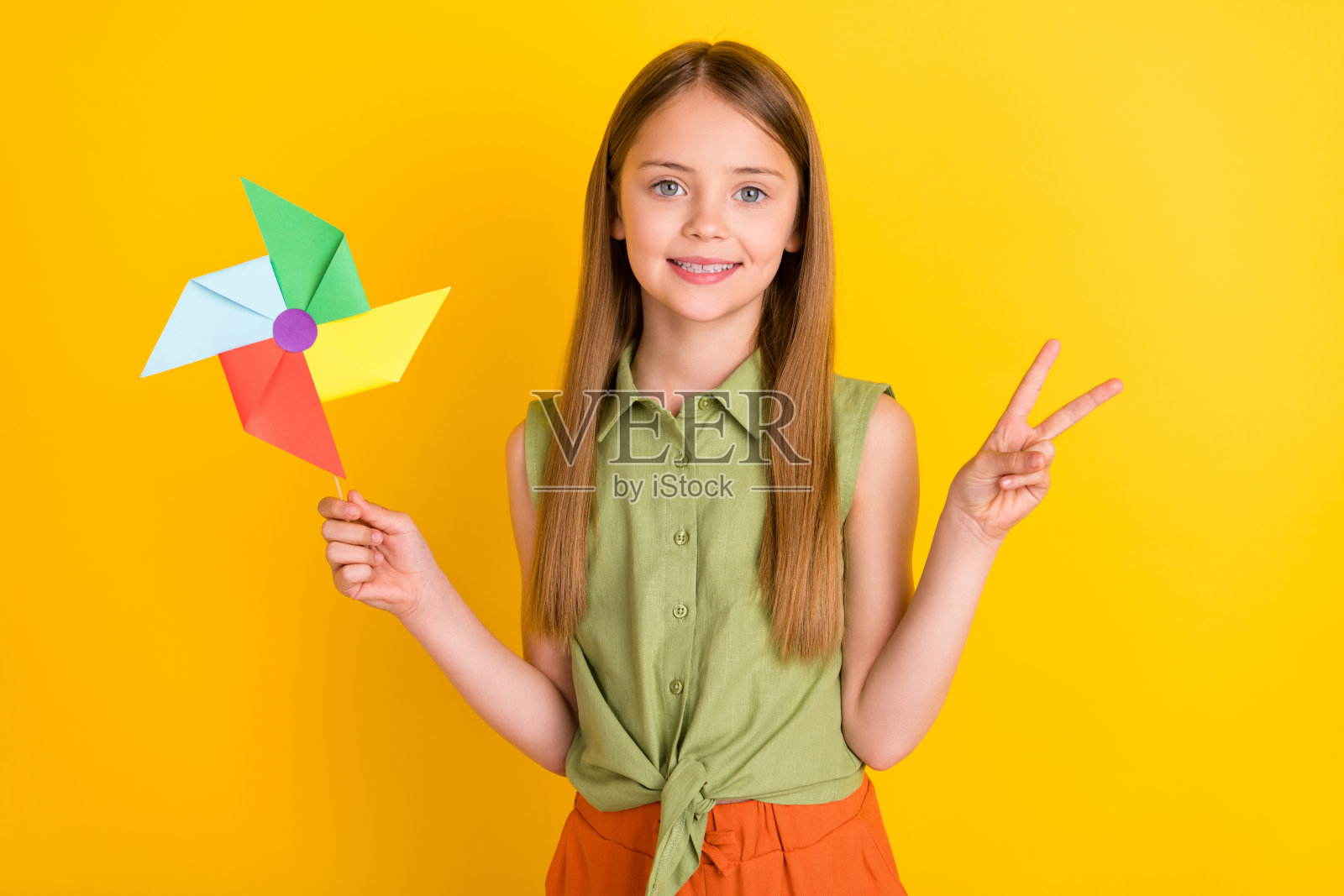 可爱的金发小女孩拿着玩具展示v-sign穿着卡其色衬衫孤立在黄色背景上照片摄影图片