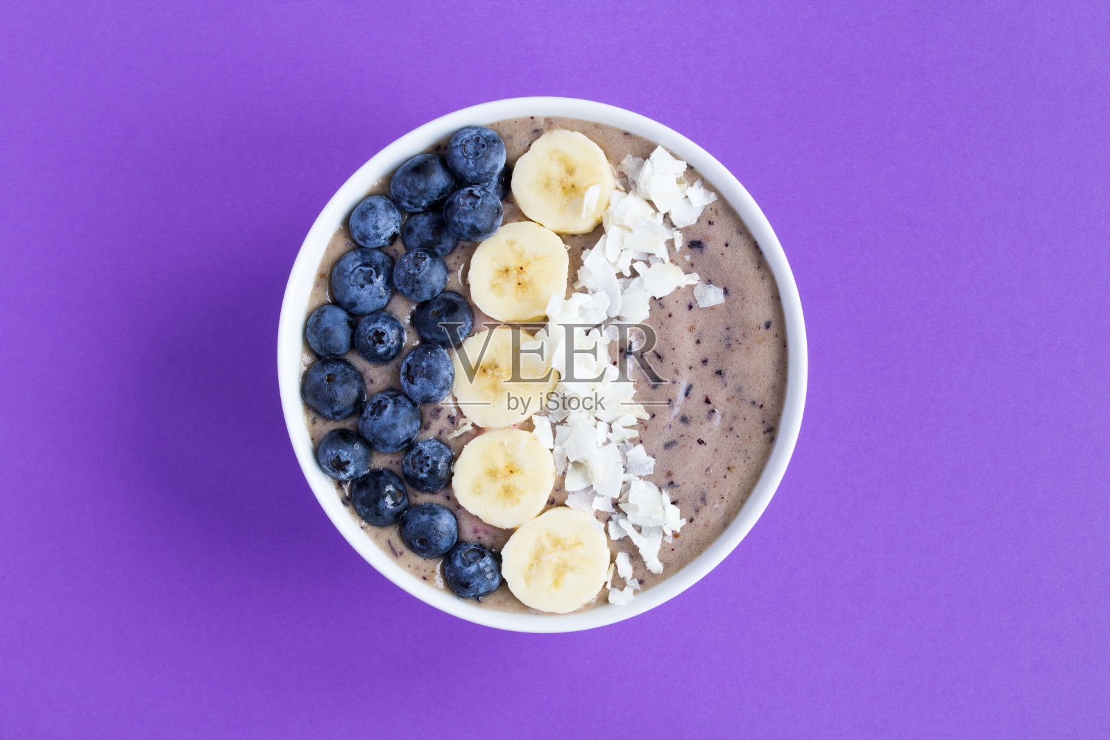 冰沙碗，紫色背景上有蓝莓，香蕉和椰子片。前视图。照片摄影图片