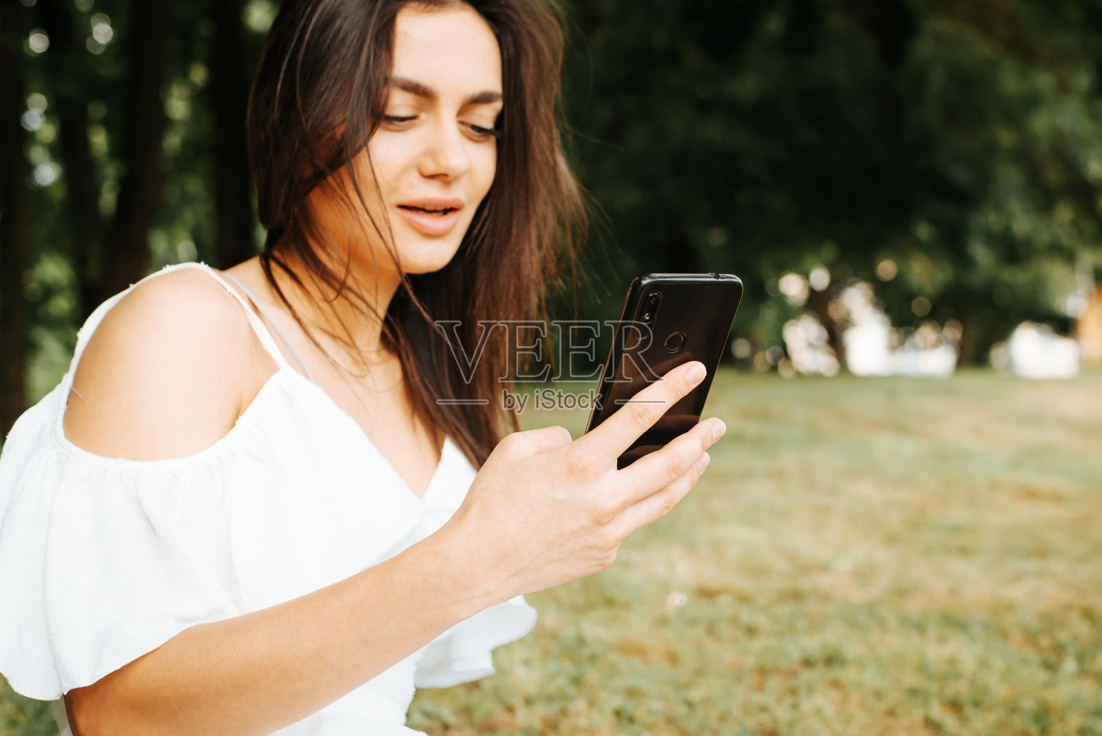 在公园户外使用手机、在社交媒体上用智能手机聊天的白色夏装年轻棕发女子，焦点对准手机照片摄影图片