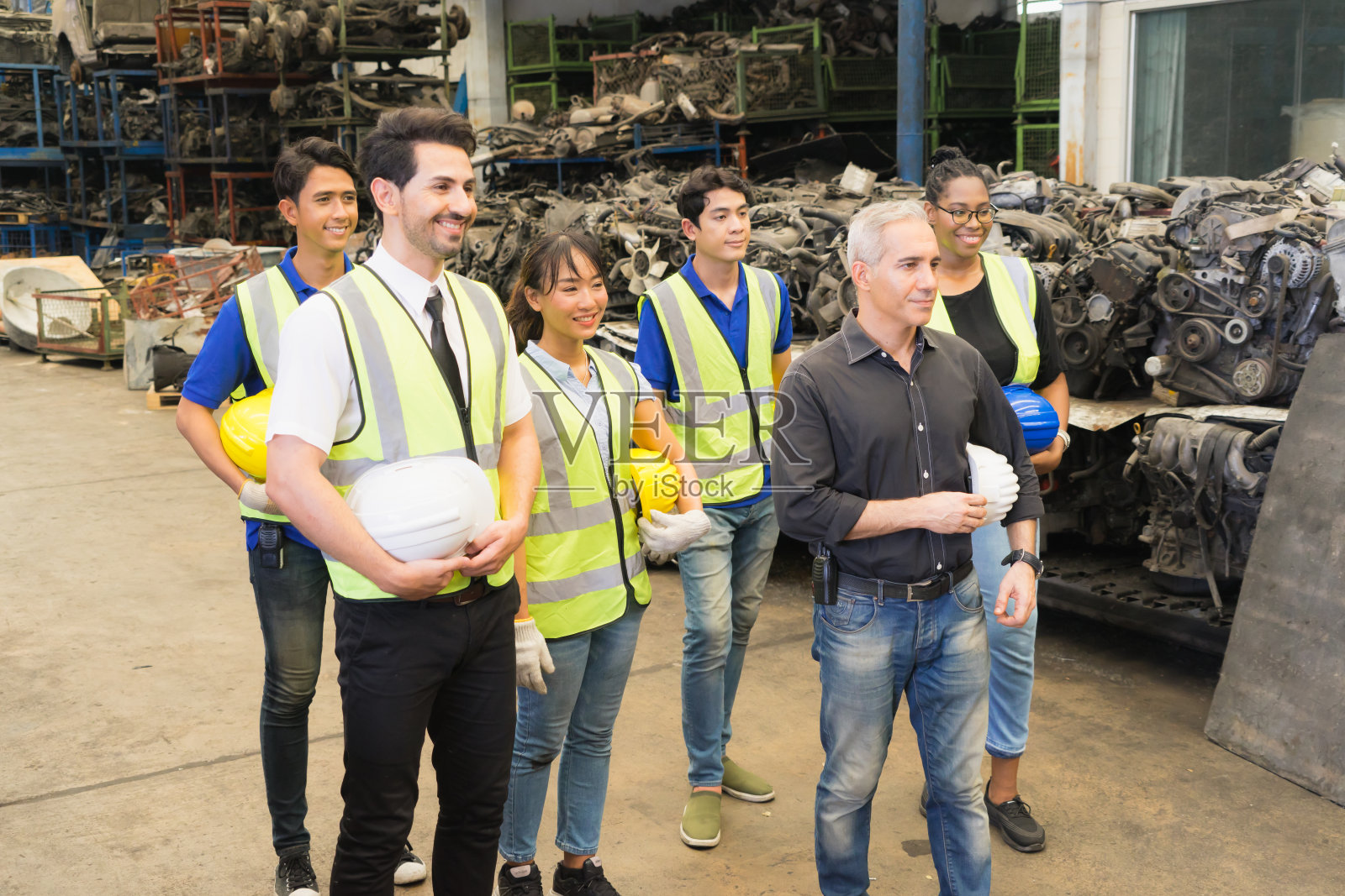 男人和女人一起工作，通过拿着头盔和看右边来显示团队合作。白人男工程师，黑人女工程师，亚裔男工程师，亚裔女工程师在工厂仓库里扮演着聪明的角色照片摄影图片