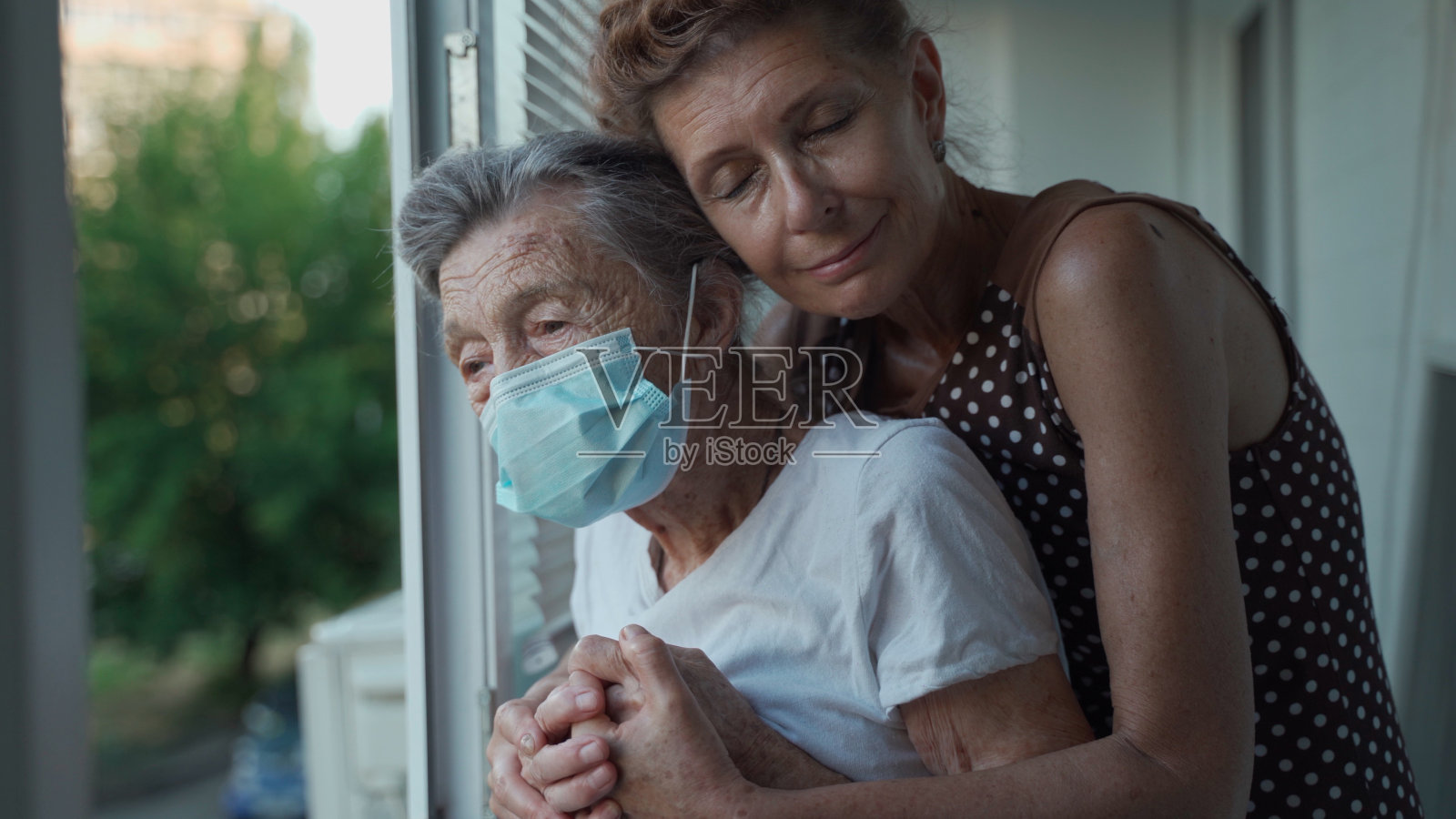 在疗养院里，戴着医用口罩的90岁老人和她成熟的女儿站在一起，看着窗外。成年的女儿探望年迈的母亲，非常爱她和支持她照片摄影图片