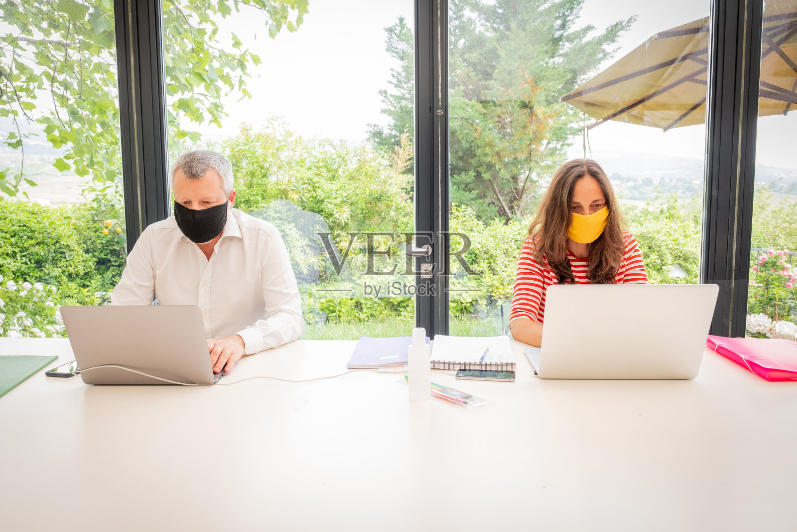 一对夫妇带着防护口罩在家里用他们的笔记本电脑工作——母亲和父亲因covid - 19大流行而在家里远程工作，进行自动隔离照片摄影图片