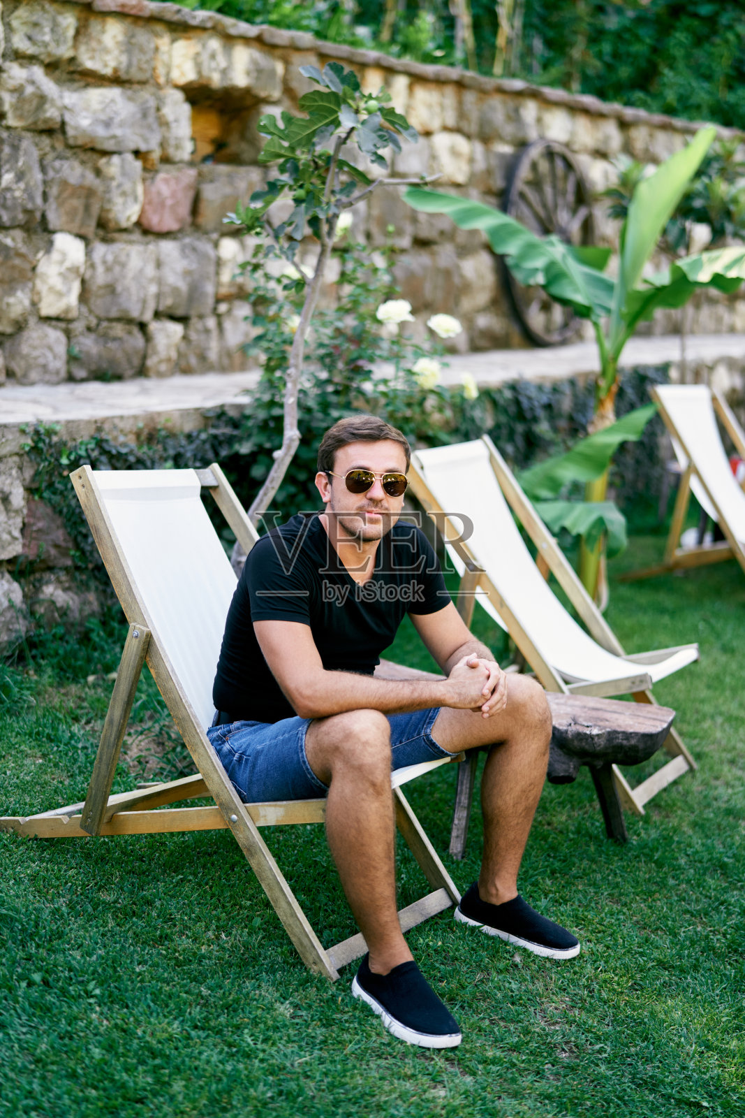 一个戴着太阳镜的男人坐在一张折叠椅上，草地靠近一堵石墙照片摄影图片