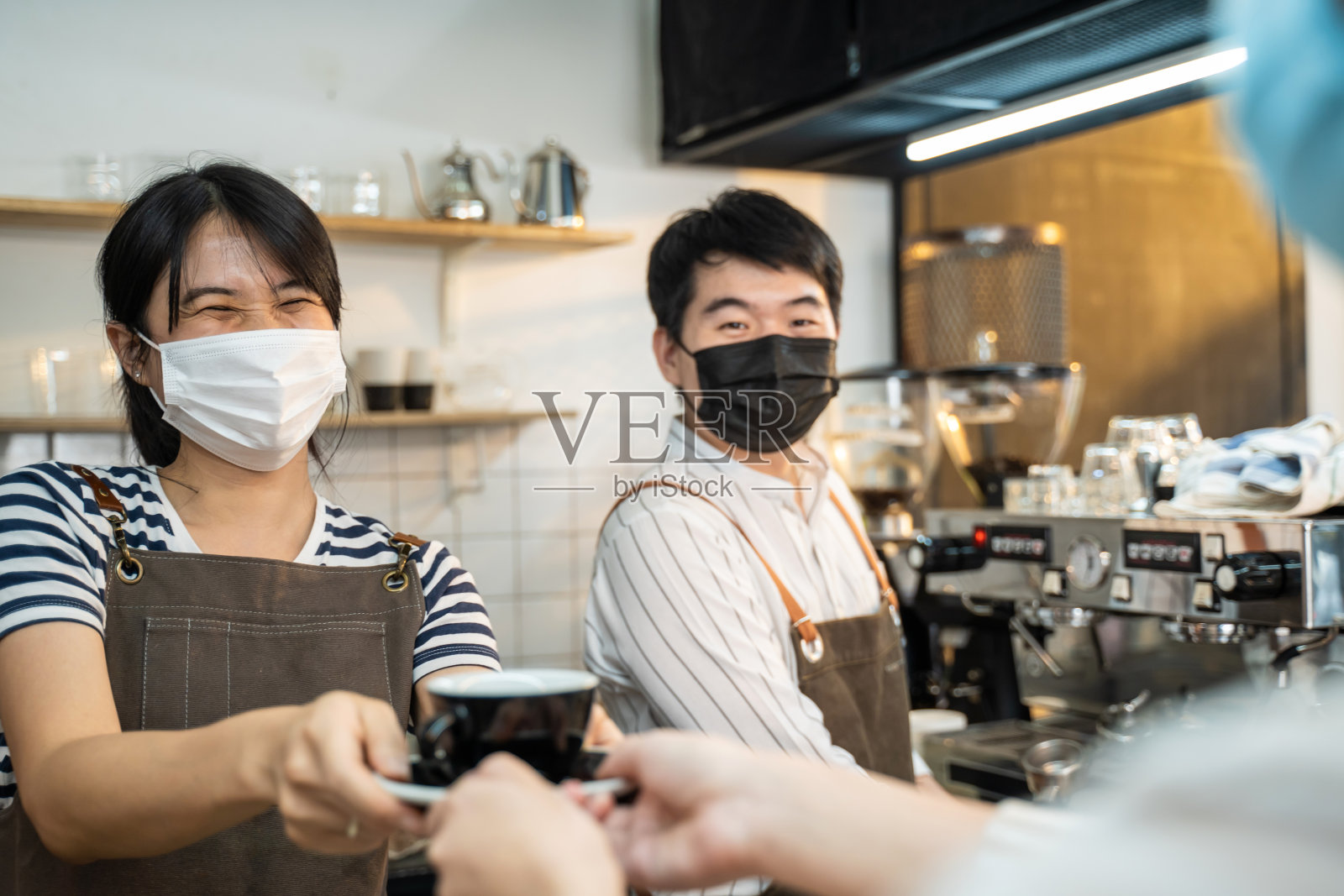 在咖啡厅，亚洲女服务员用双手将一杯咖啡递给顾客。穿着围裙的女老板咖啡师因新冠肺炎疫情戴口罩，在餐厅服务的职业服务年轻女孩。照片摄影图片