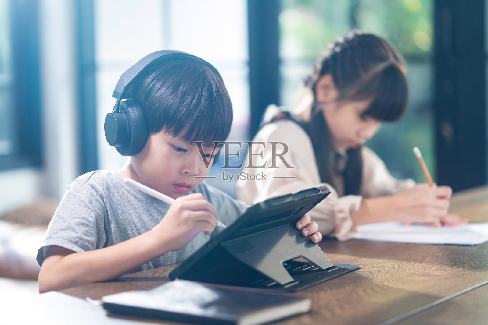 亚洲小孩子在家里客厅的桌子上自学。可爱男孩使用平板电脑和耳机进行在线学习，女孩做作业时在纸上写笔记。教育和科技理念。照片摄影图片