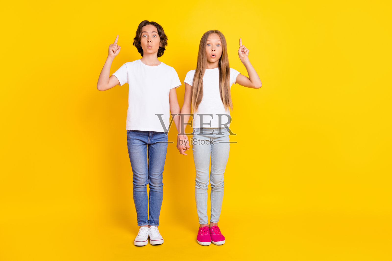 两个迷人的惊奇的孩子的全身尺寸的观点，手牵手，食指指向孤立在明亮的黄色背景照片摄影图片