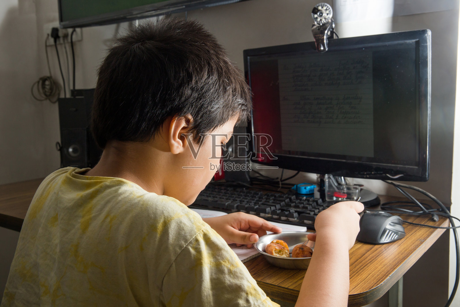 一名8岁的印度男孩因为冠状病毒而被锁在家里，在带摄像头的台式电脑上学习在线课程，一边上课一边吃早餐照片摄影图片
