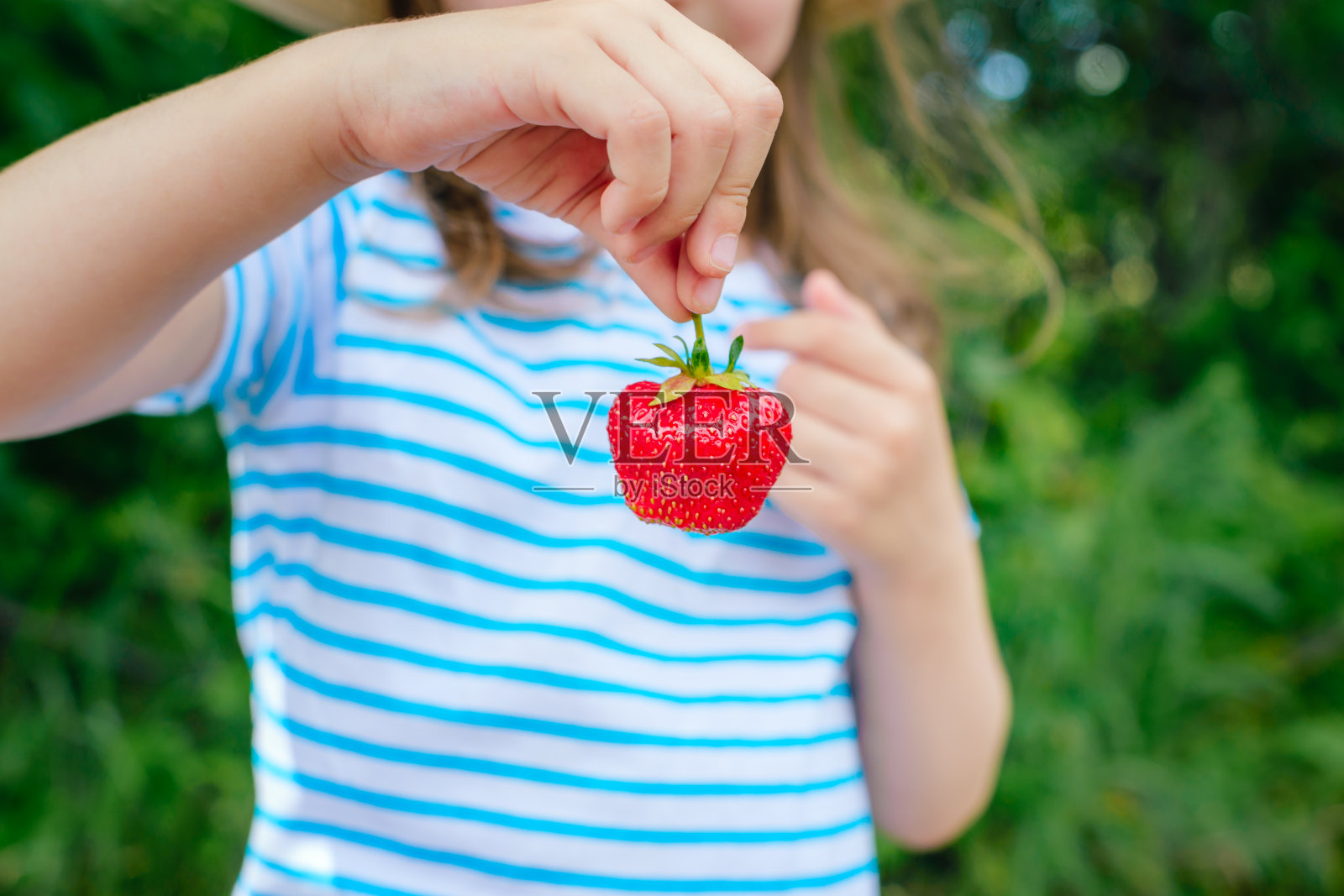 快乐的小女孩咬着一颗大草莓。照片摄影图片