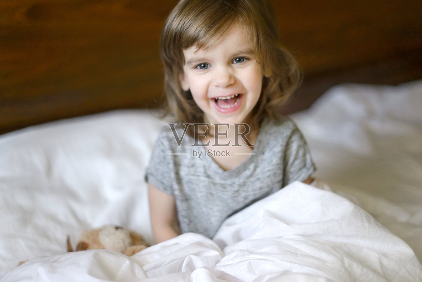 活泼可爱的宝宝醒来时躺在床上，躺着她的泰迪熊玩具，柔软的白色床单。儿童节。世界微笑日。正确的开始早晨。用微笑开始新的一天。照片摄影图片