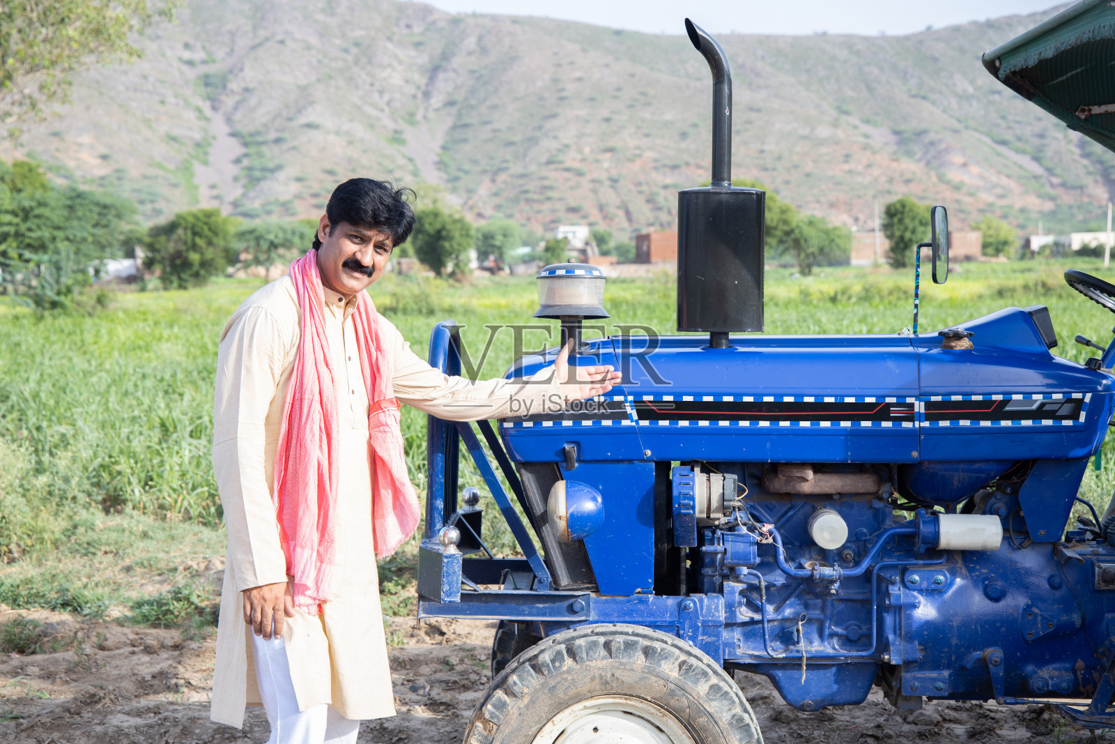 年轻快乐的印度农民展示他的蓝色拖拉机站在农田。一名男子穿着传统的无领短衫，留着胡子，微笑着看着镜头。印度农村的概念。照片摄影图片