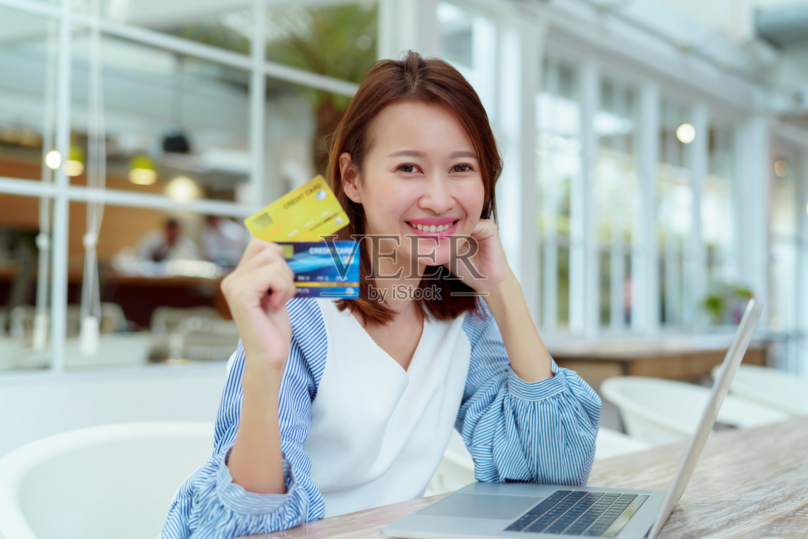 一位身穿白衬衫的亚洲美女正坐在电脑前，手里拿着信用卡准备网购。她在面包店里看起来很开心。照片摄影图片