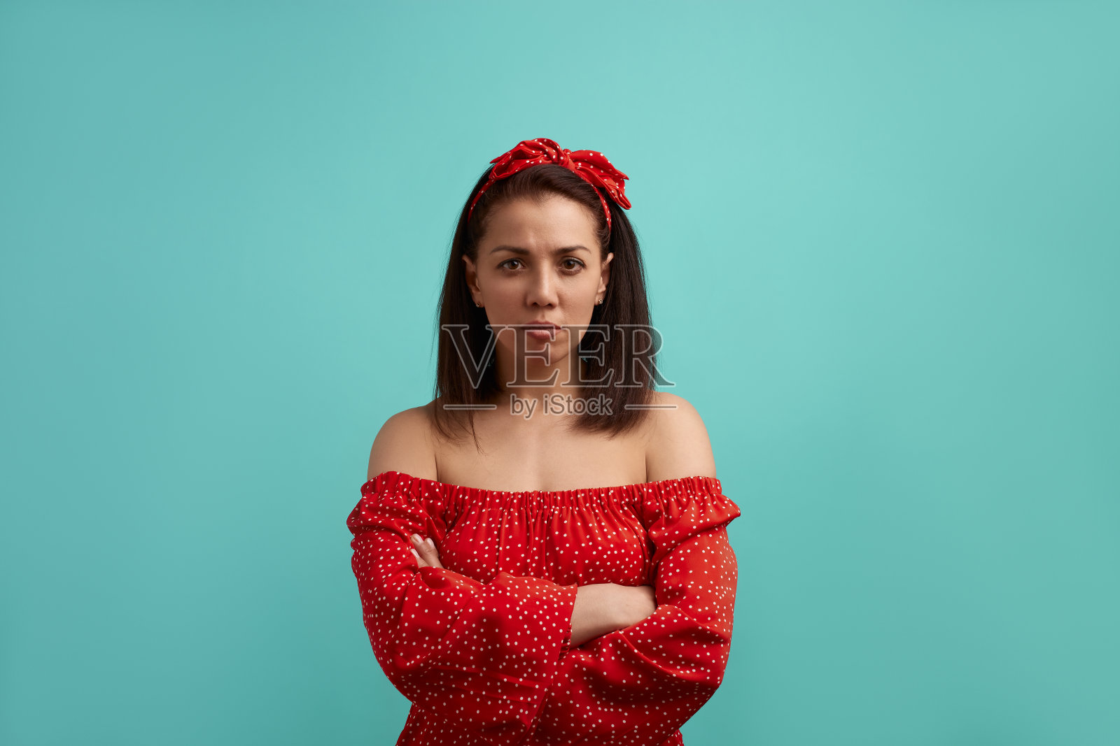 自信而冷静的欧洲女性，深褐色的红色露肩礼服，带着发带站直看着镜头，双臂交叉在胸前，孤立在蓝绿色的背景上。照片摄影图片