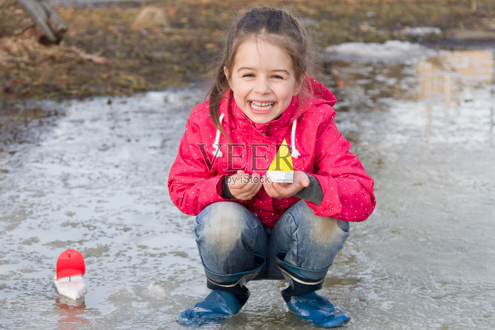 快乐可爱的小女孩穿着雨靴在春天的小溪里玩玩具船照片摄影图片