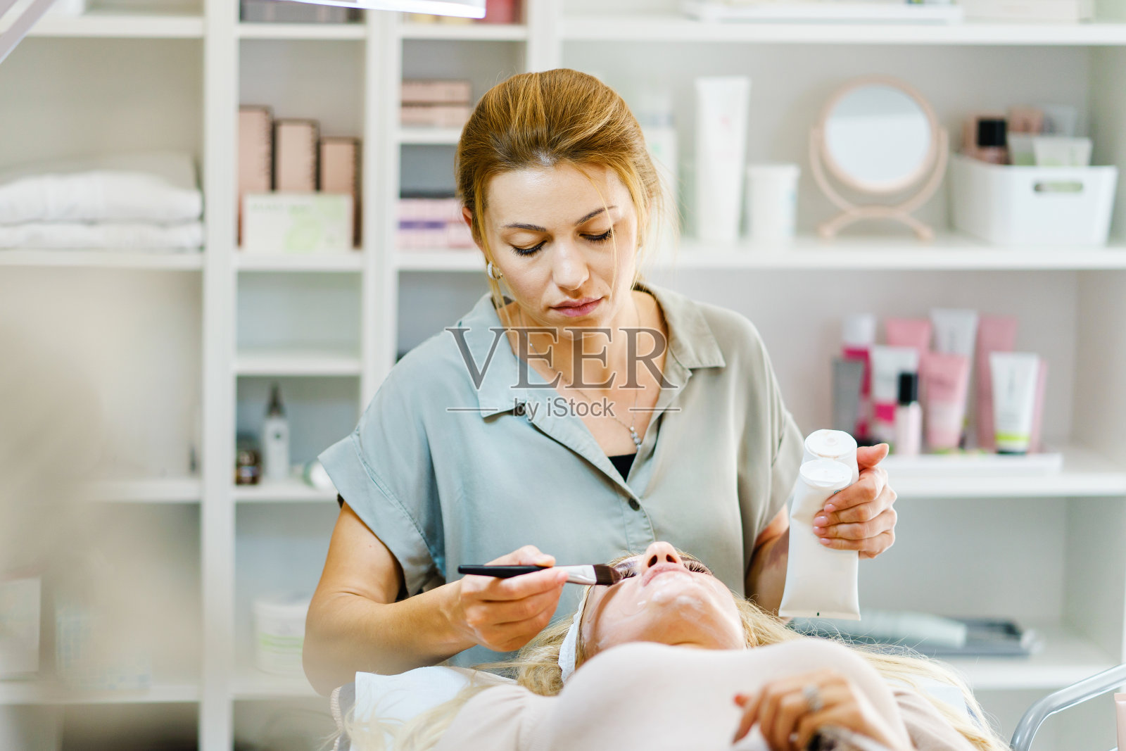 美丽快乐的白人女性微笑着在水疗中心给客户做面部护理-美容师，美容师在美容院用刷子涂面膜-保健概念复制空间照片摄影图片