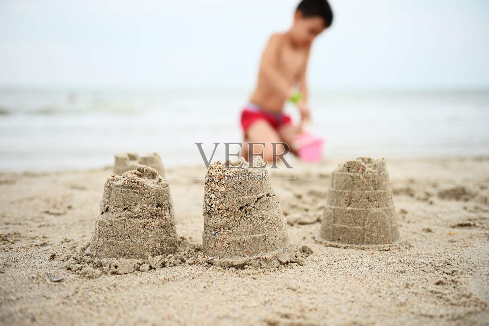 沙堡的背景，模糊男孩穿红色泳裤。一个十几岁的男孩在沙滩上堆砌人物，享受着积极的暑假。暑假的概念照片摄影图片