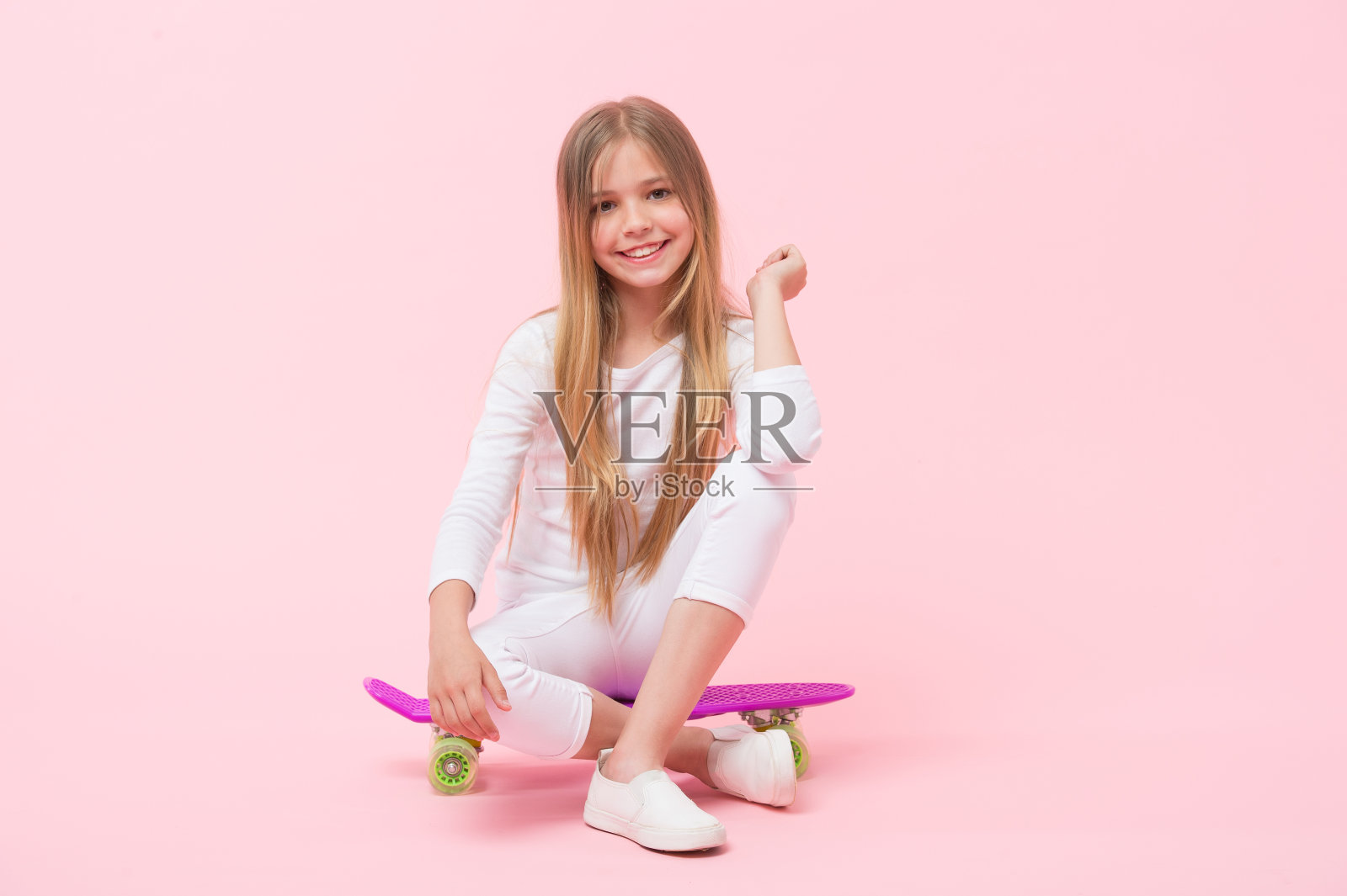 享受硬币板上的共鸣。可爱的小孩坐在粉红色背景的硬币板甲板上。可爱的女孩溜冰与紫罗兰便士板。小滑板与便士滑板照片摄影图片