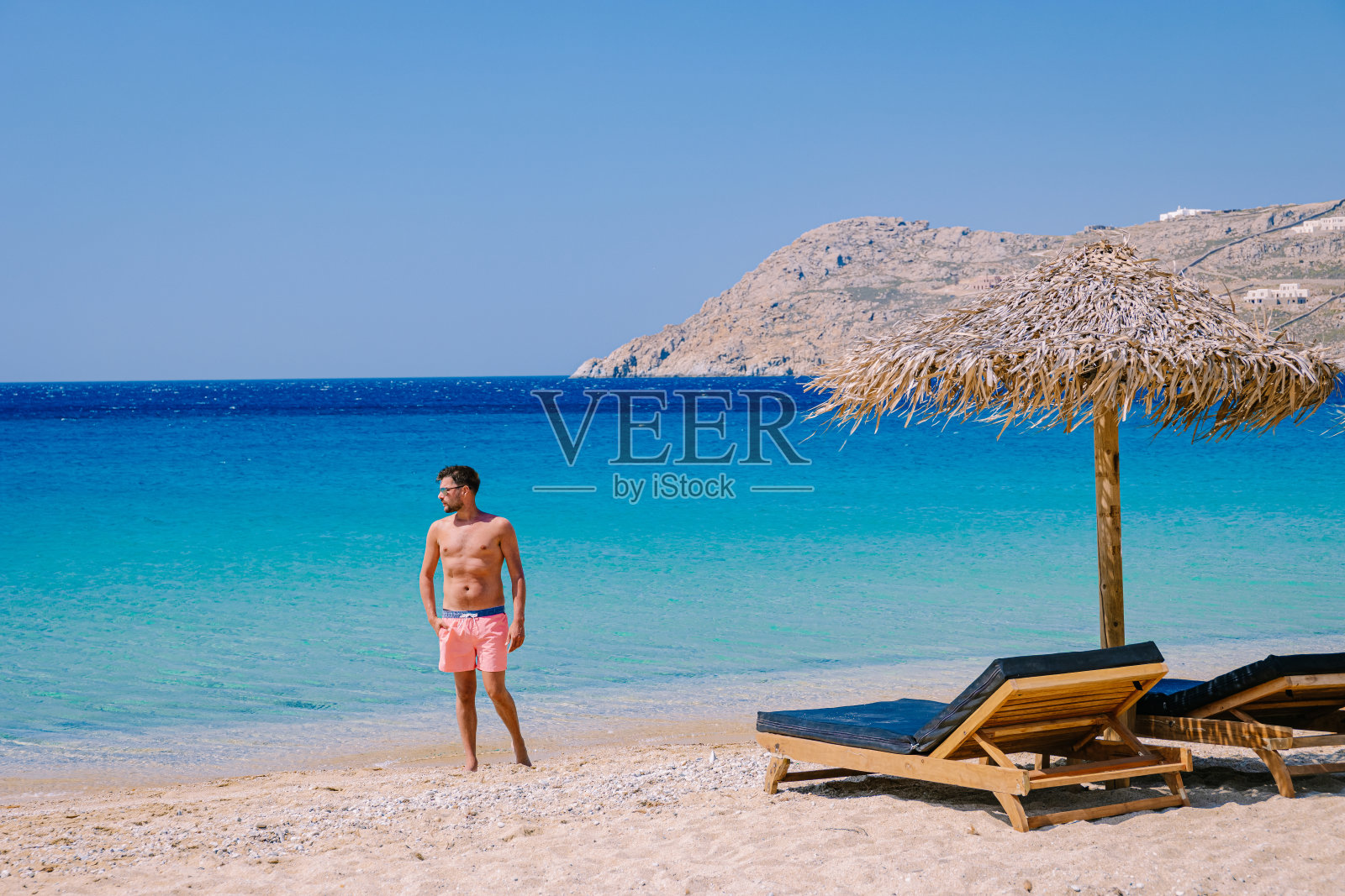 年轻的小伙子在米科诺斯海滩，米科诺斯海滩，夏天的米科诺斯海滩配有伞和豪华沙滩椅床，蓝色的海洋在希腊米科诺斯的埃利亚海滩照片摄影图片