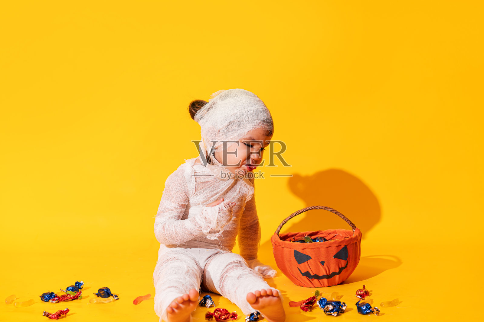 穿着木乃伊服装的孩子在黄色背景下吃着篮子里南瓜形状的糖果照片摄影图片