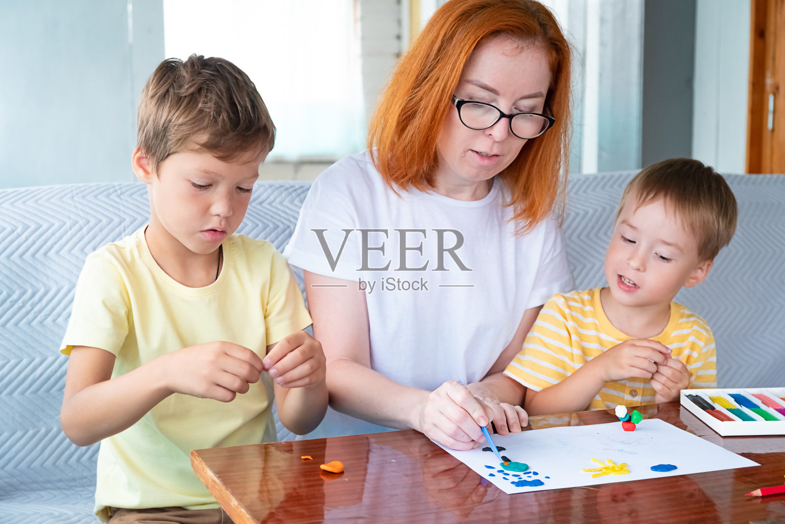 两个白种男孩和戴着眼镜的红发女人在幼儿园或家里玩彩色橡皮泥。母亲和儿子或保姆都从事早期发育，为上学做准备照片摄影图片