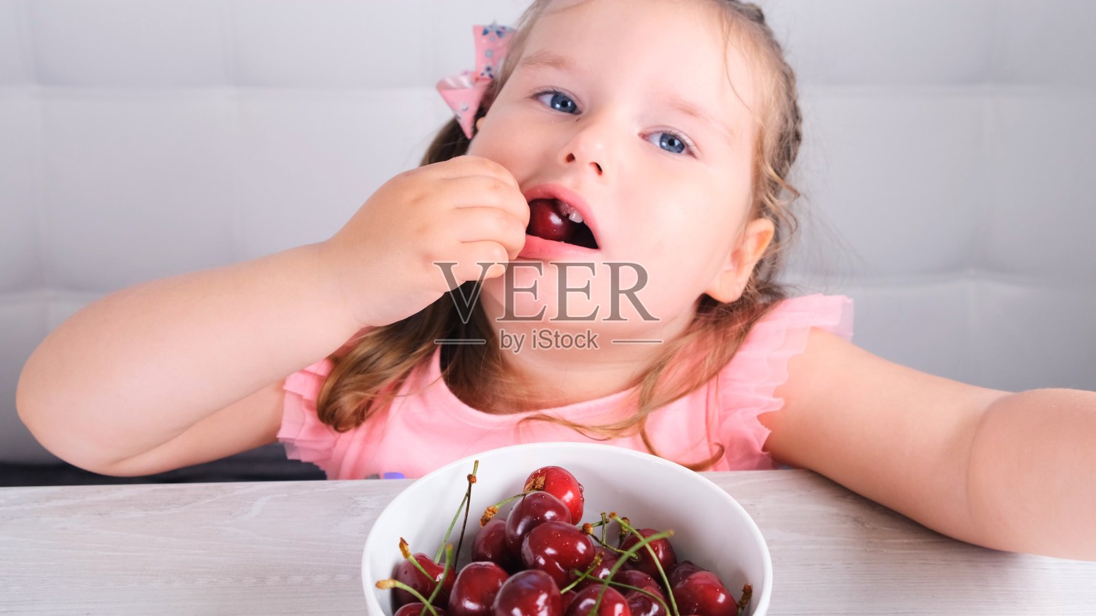 可爱的金发小女孩坐在一张轻便的木桌旁，桌上放着一盘樱桃，吃着红色的浆果。健康饮食照片摄影图片