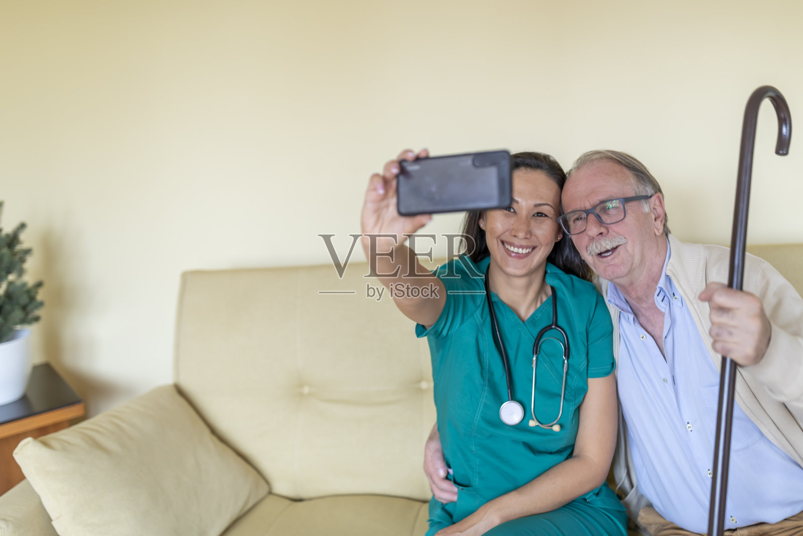 穿着绿色制服的年轻护士正在和她的老年病人在家访中拍照。照片摄影图片