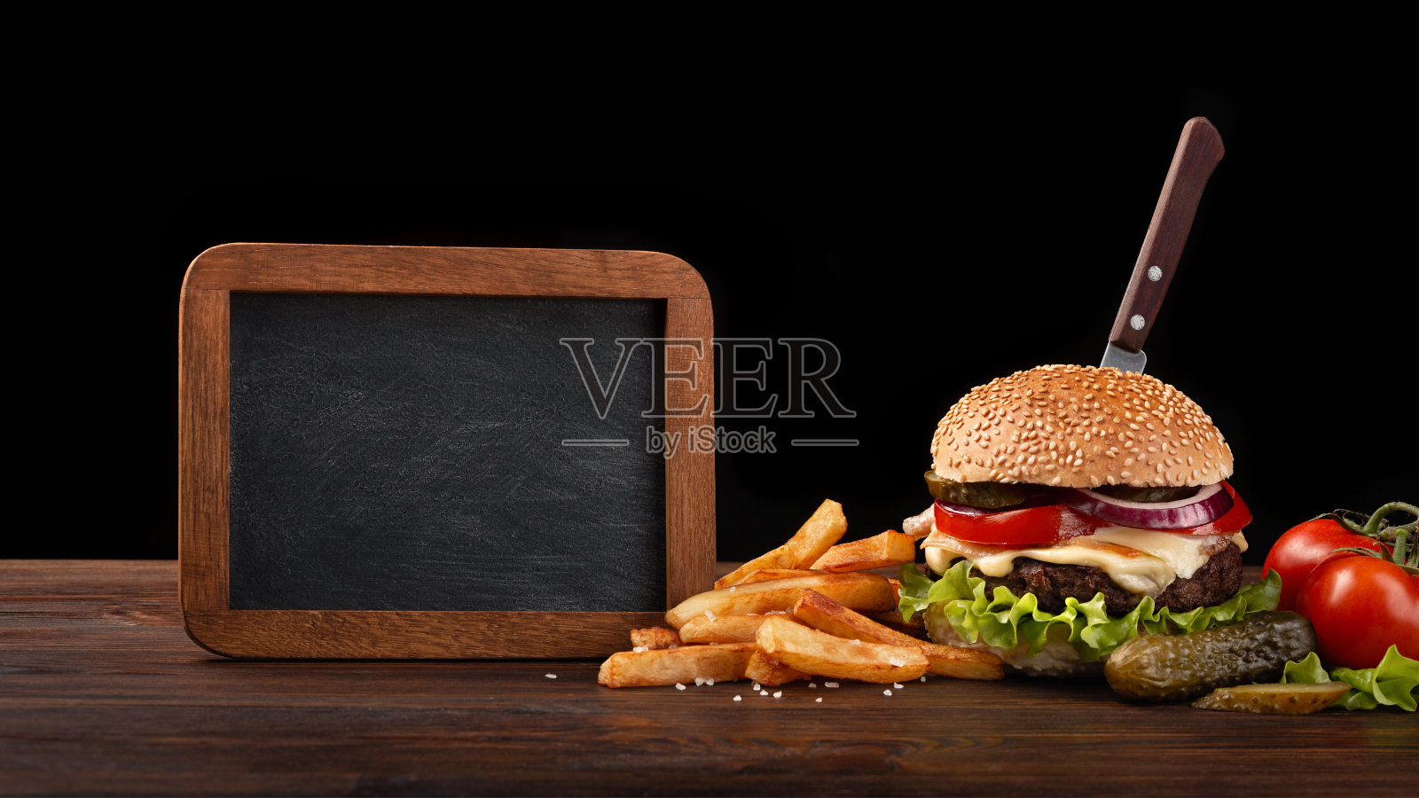 自制汉堡的特写，牛肉，西红柿，生菜，奶酪，薯条和黑板在木桌上。一把刀插在汉堡里照片摄影图片