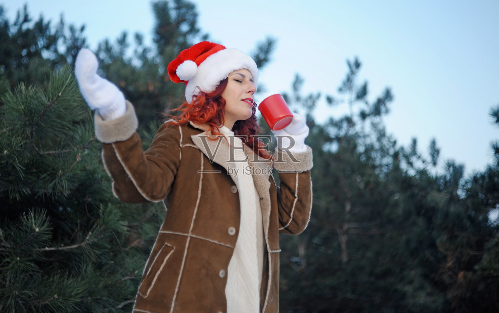 戴着圣诞帽的红头发女人在户外喝热咖啡。冬天的假期。照片摄影图片