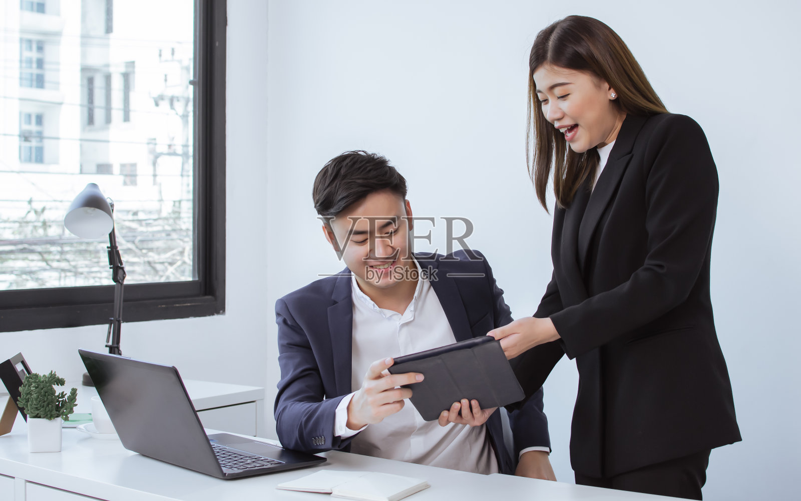 亚洲年轻英俊的商务男子和漂亮的秘书穿着正式的西装，微笑着在办公室里，一个女人用平板电脑向老板介绍她的项目。照片摄影图片
