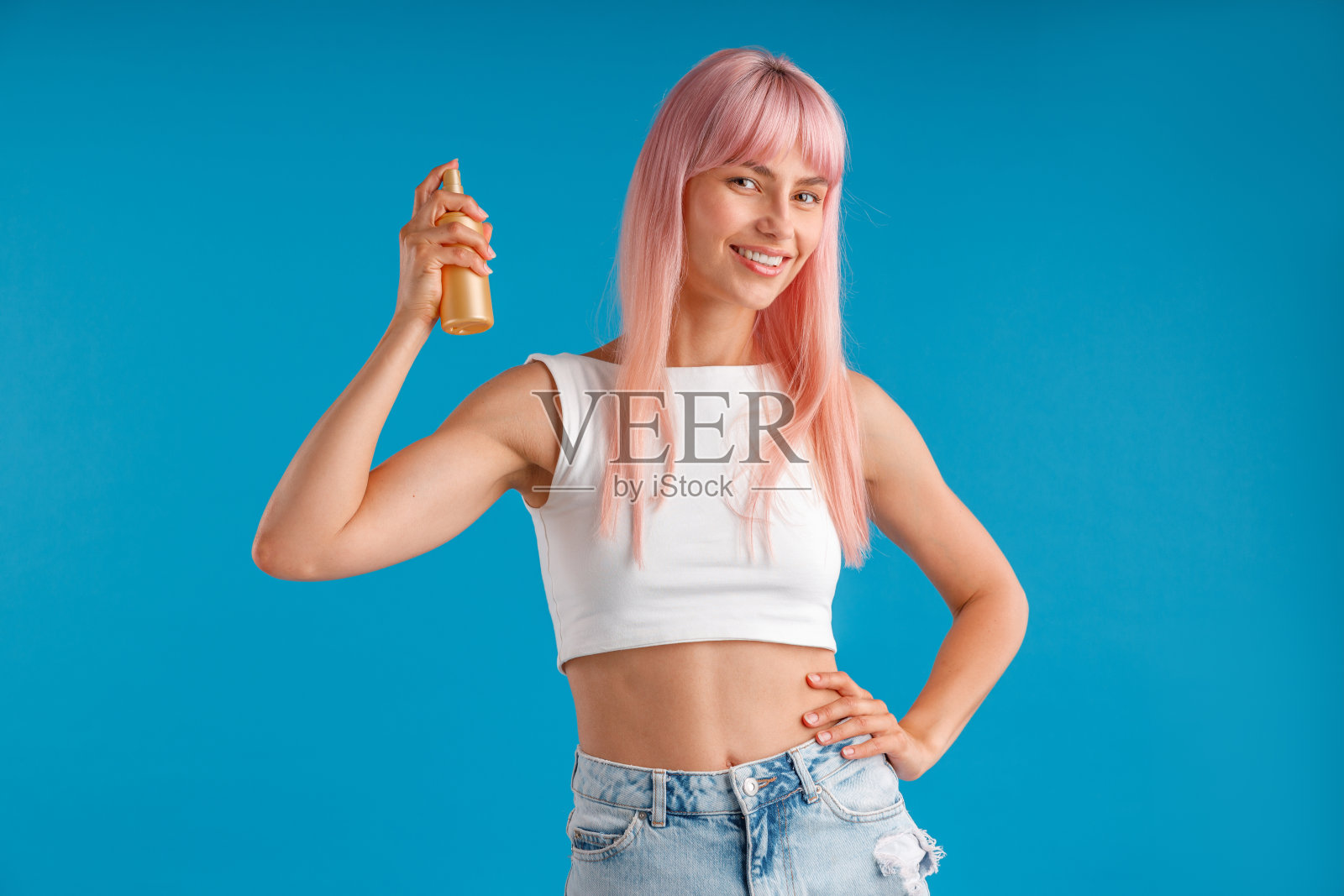 快乐的年轻女子粉红色的头发微笑，拿着一瓶护发素，保湿喷雾，孤立地站在蓝色的工作室背景照片摄影图片
