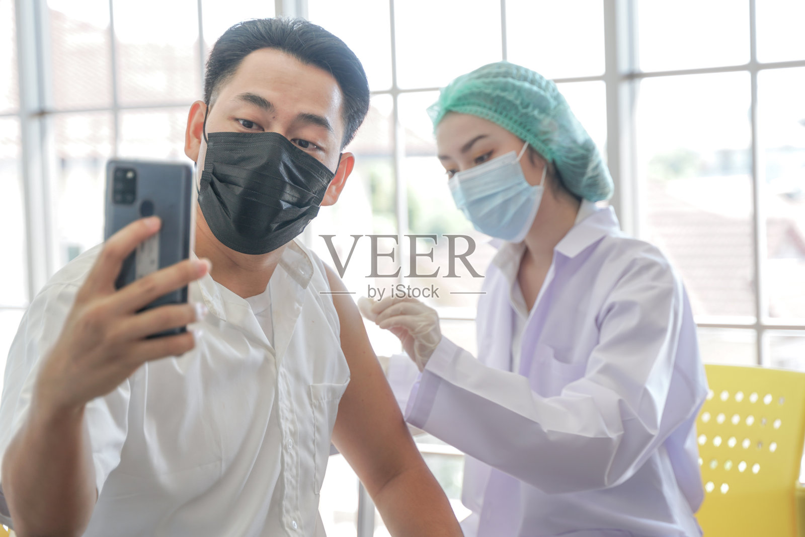 图为一名亚洲男子在诊所或医院接种新冠疫苗时通过手机自拍，护手注射疫苗以获得免疫力以保护病毒。人们戴着防护口罩。照片摄影图片